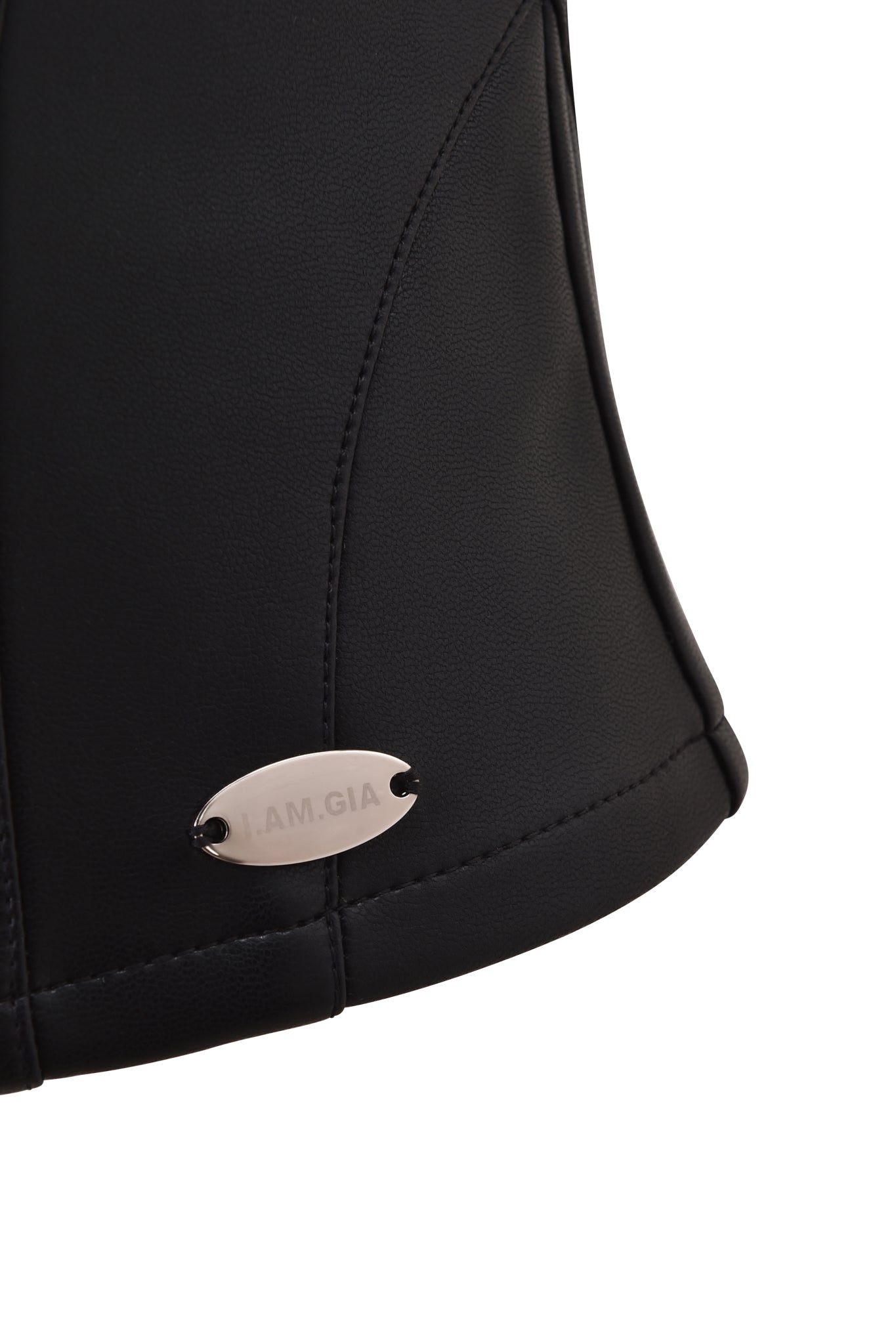Close-up of the lower side of the TEMPEST BUSTIER TOP - BLACK by I.AM.GIA, featuring visible stitching and a small, oval metallic tag engraved with the brand name attached to the black PU fabric.