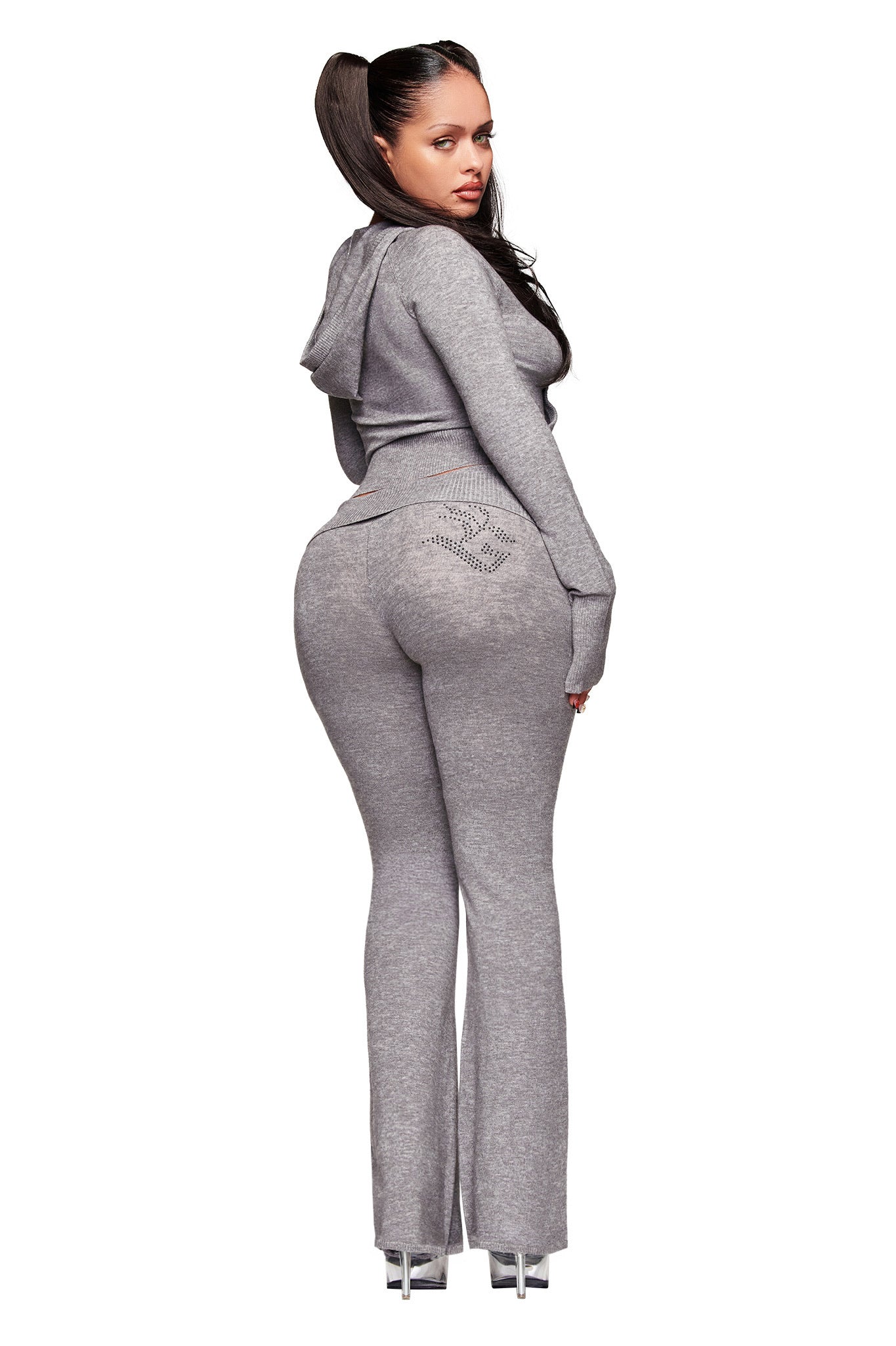 A woman with long brown hair in a high ponytail models the I.AM.GIA JESSA PANT - GREY tracksuit, standing on high heels and looking over her shoulder against a plain white background.