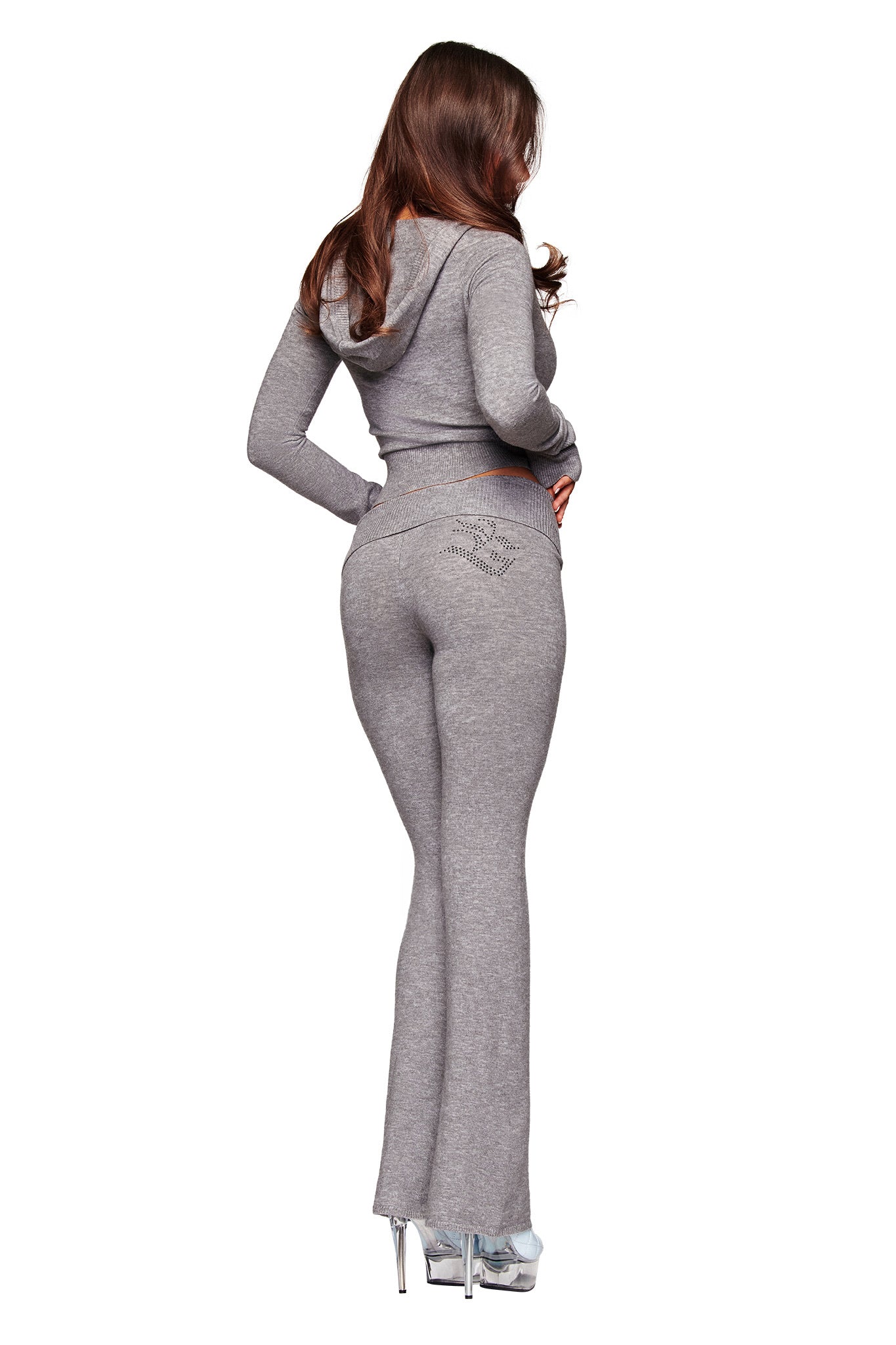A woman with long brown hair stands with her back to the camera against a white background, wearing the I.AM.GIA JESSA PANT in grey—a fitted hooded tracksuit with flared legs—paired with silver high heels.