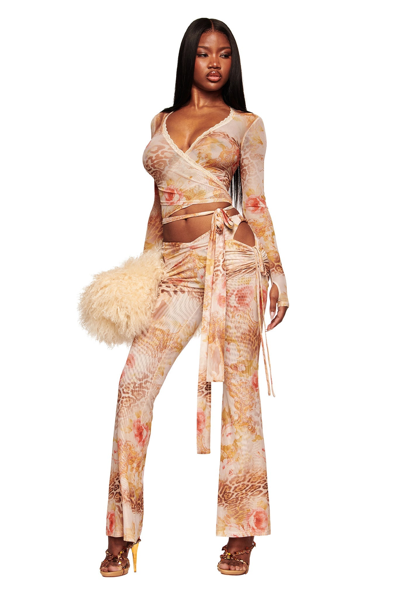 A woman wears the I.AM.GIA ERISKA PANT - DESERT ROSE, featuring a pastel abstract print, with a fitted long-sleeve wrap top. She holds a fluffy beige handbag and wears heeled sandals, posing confidently against a white background.