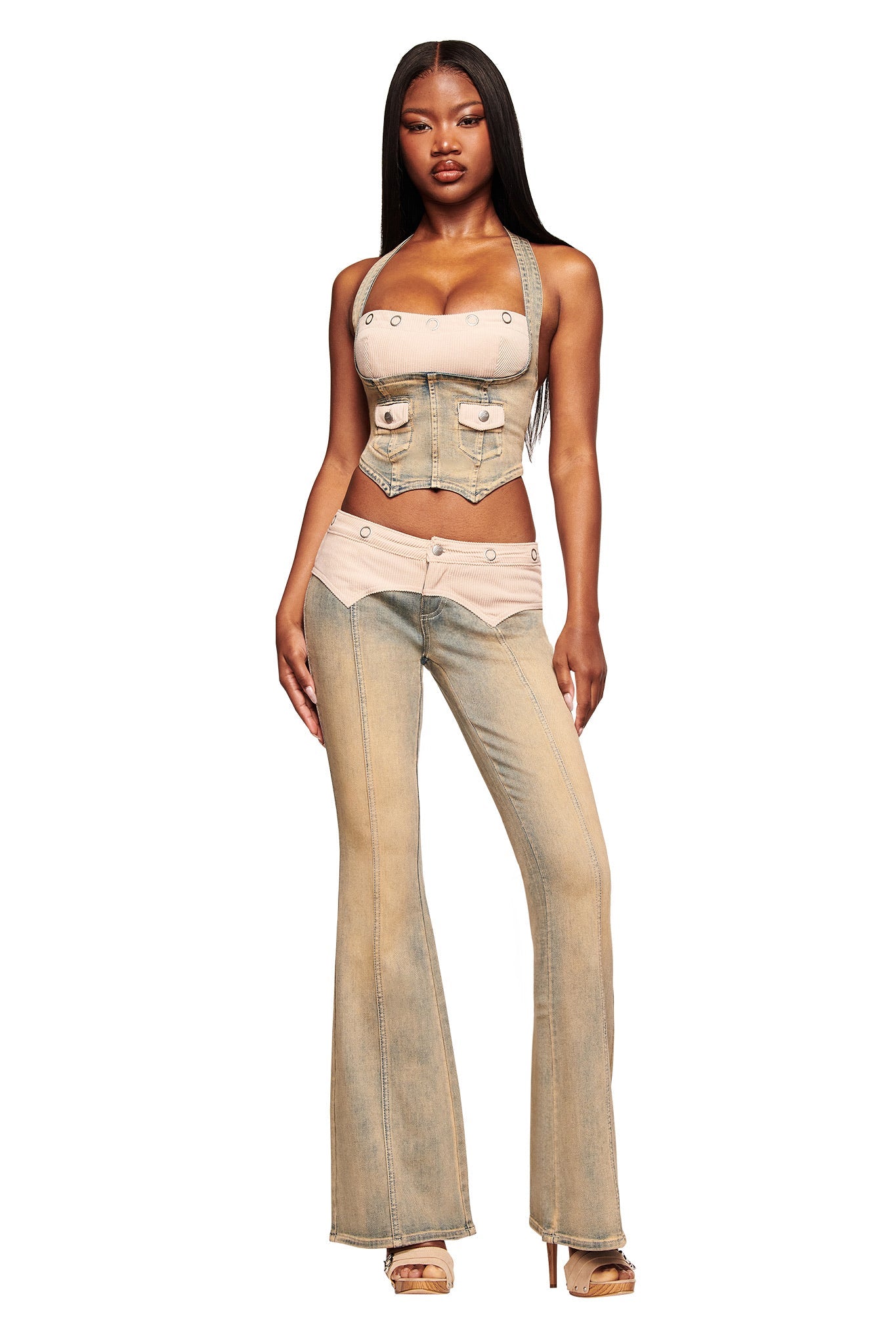 A woman stands confidently in the I.AM.GIA TOMMIE JEAN - ANTIQUE DENIM outfit, featuring a corset-style top and color-blocked flared pants with metallic accents and beige platform heels, set against a white background.