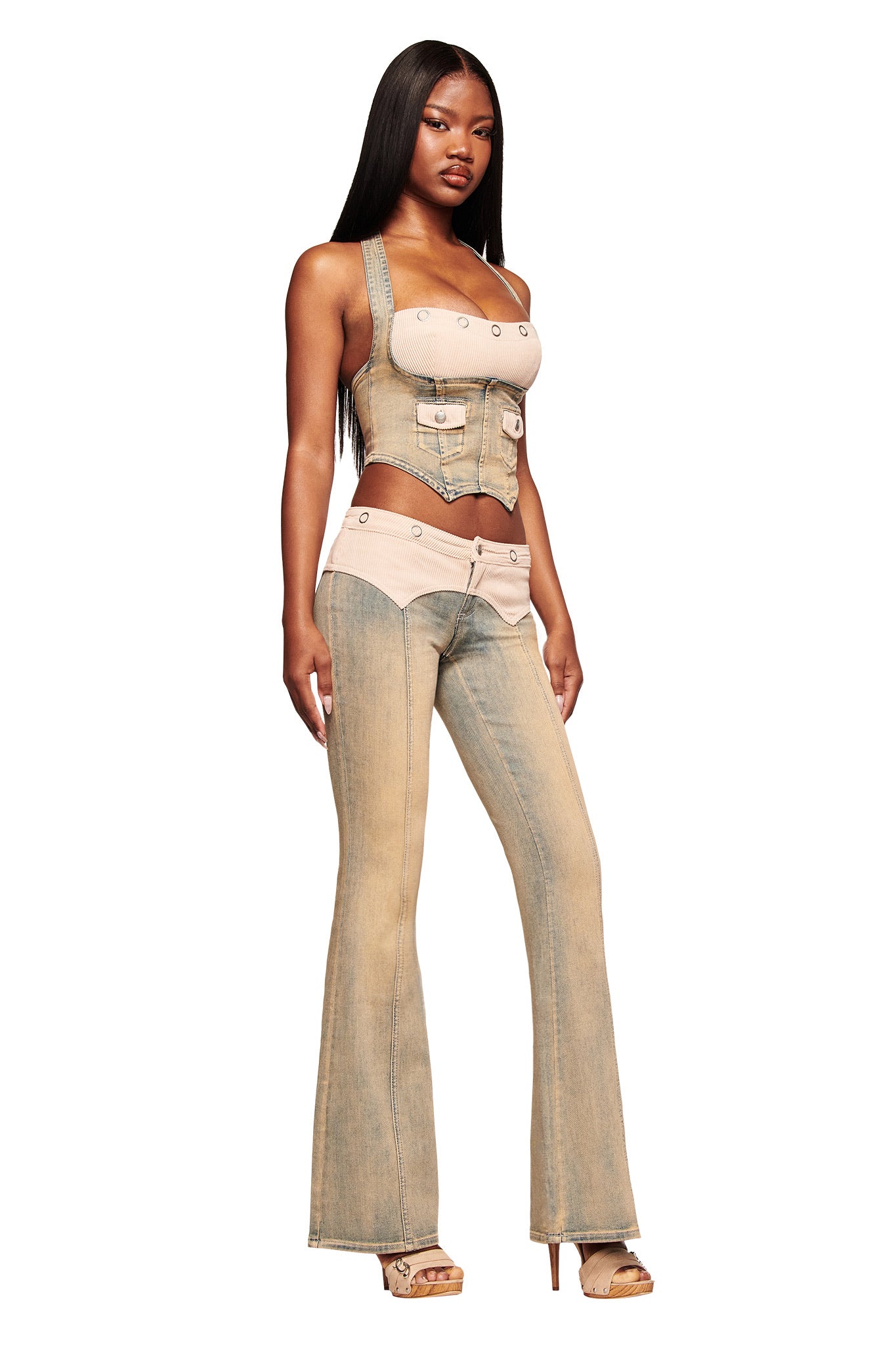 A woman with long straight hair wears the I.AM.GIA TOMMIE JEAN - ANTIQUE DENIM corset top and matching mid-rise flared pants with beige accents, paired with high platform sandals, standing against a white background.