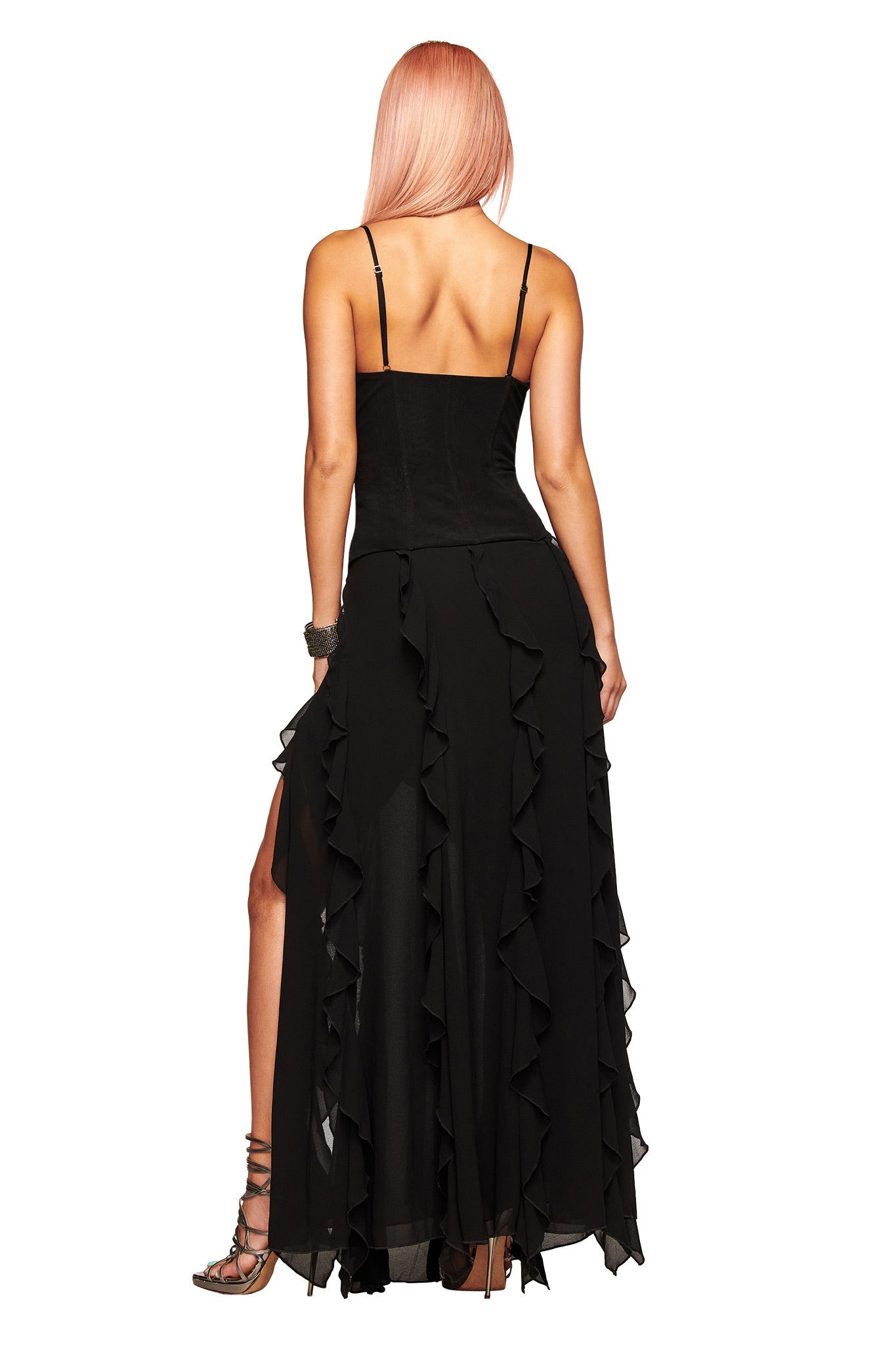 A woman with pink hair faces away wearing the I.AM.GIA MARLETTE 2.0 MAXI DRESS in black, featuring spaghetti straps and ruffled layers. She pairs the dress with sandals and a bracelet, her left leg slightly bent.