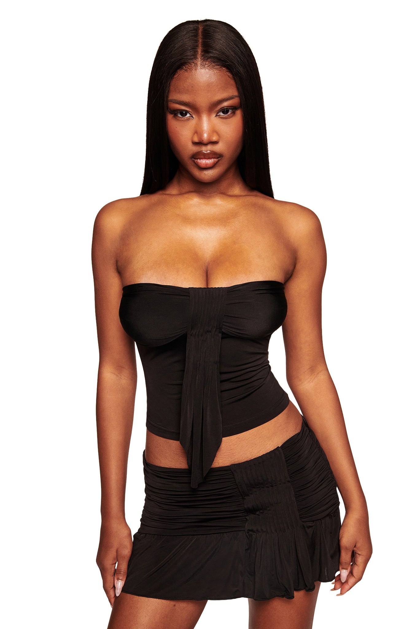 A woman with long straight hair models the I.AM.GIA ELYSIA MINI SKIRT in black, paired with a strapless black top, standing confidently against a white background.