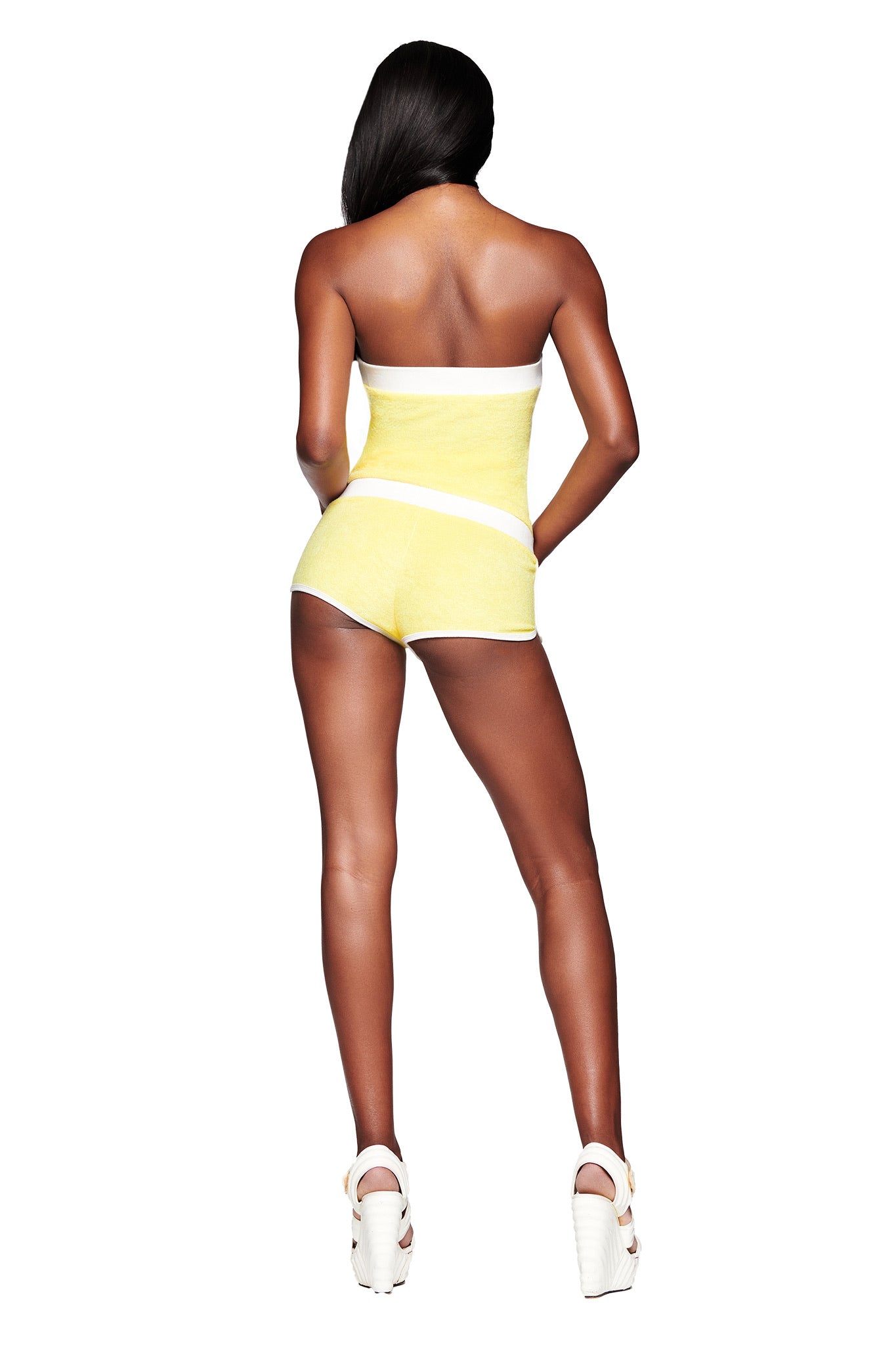 A woman faces away, wearing the I.AM.GIA GOLDIE ROMPER in yellow—featuring a pastel yellow and white boob tube top and dolphin hem shorts—with white platform heels, against a plain white background.