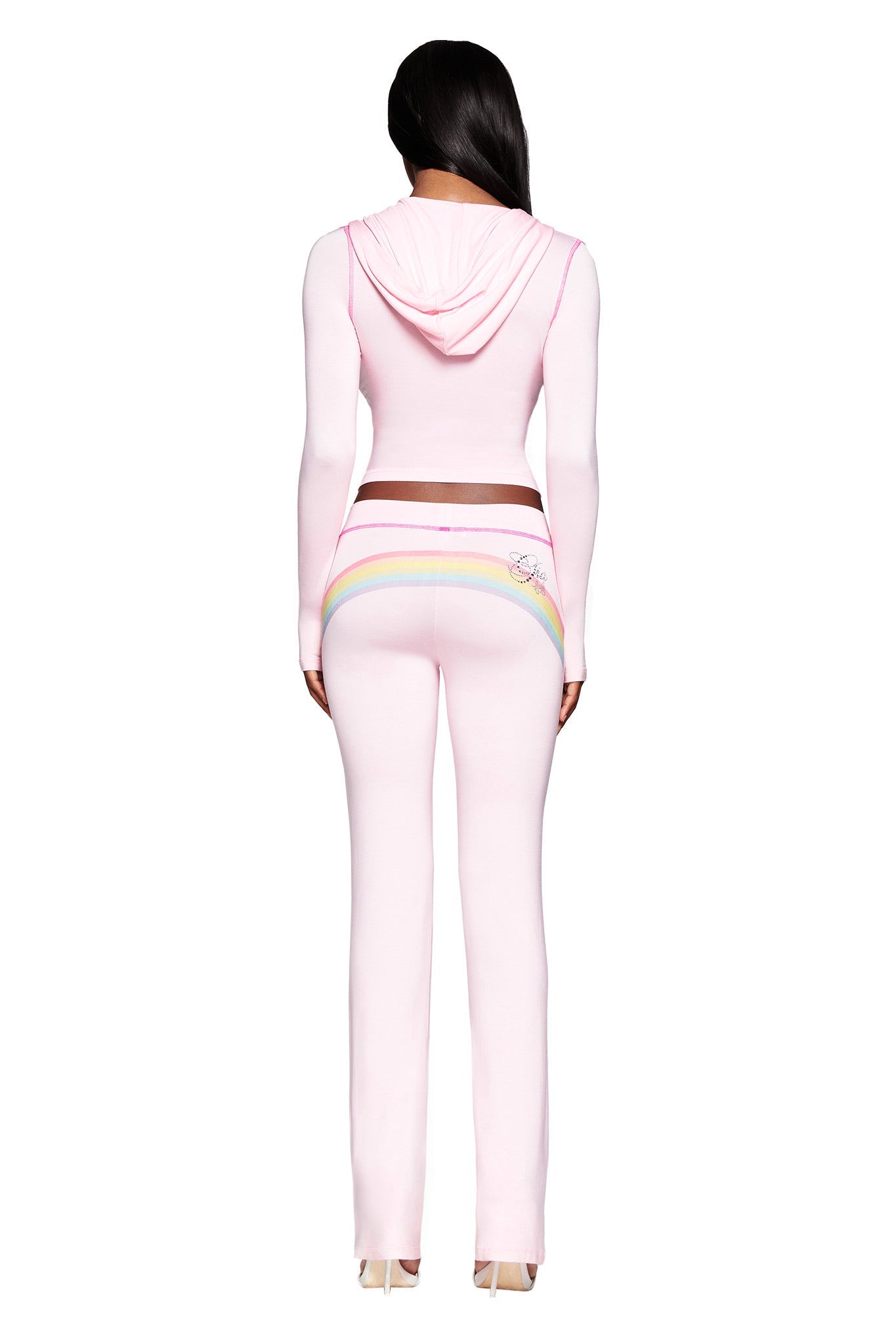 A woman faces away, wearing the I.AM.GIA FAFI BLARE HOODIE in baby pink with matching pants, featuring a rainbow design, diamanté accents, and script on the back, all set against a white background.