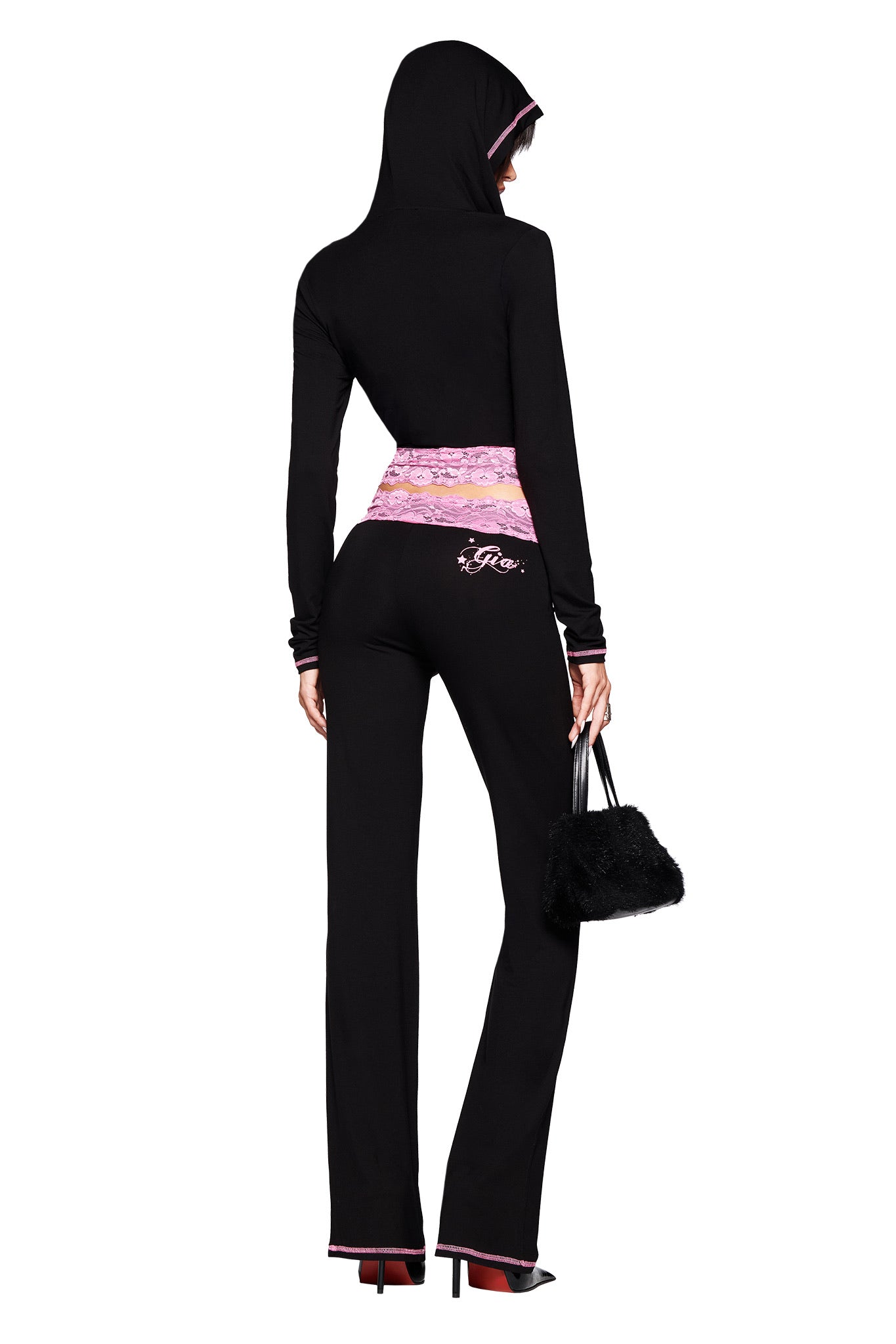 A person stands facing away, wearing the I.AM.GIA TARSHA JACKET in black/pink with a body-con fit and pink waistband accents, paired with a small black purse and high heels.