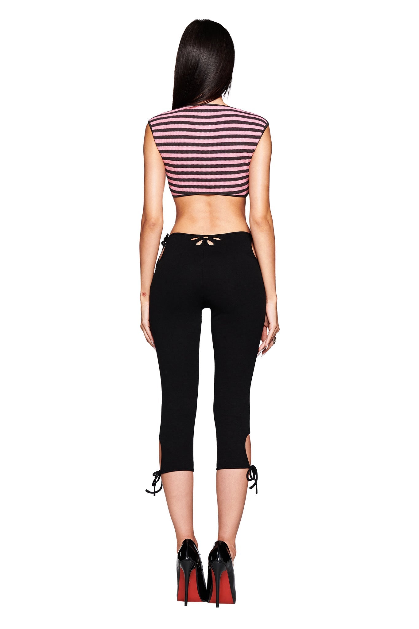 A woman with long dark hair faces away, wearing I.AM.GIA’s YUNA CAPRI - BLACK mid-rise body-con leggings featuring floral cutout and bow details, paired with a maroon and white striped cropped tee and black heels with red soles.