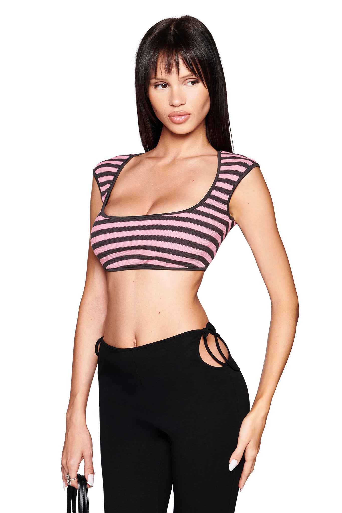 A woman with long straight hair wears the I.AM.GIA RICA TOP in pink and grey—a striped super crop tank with cap sleeves—paired with black high-waisted pants with side cutouts, holding a small black item against a white background.