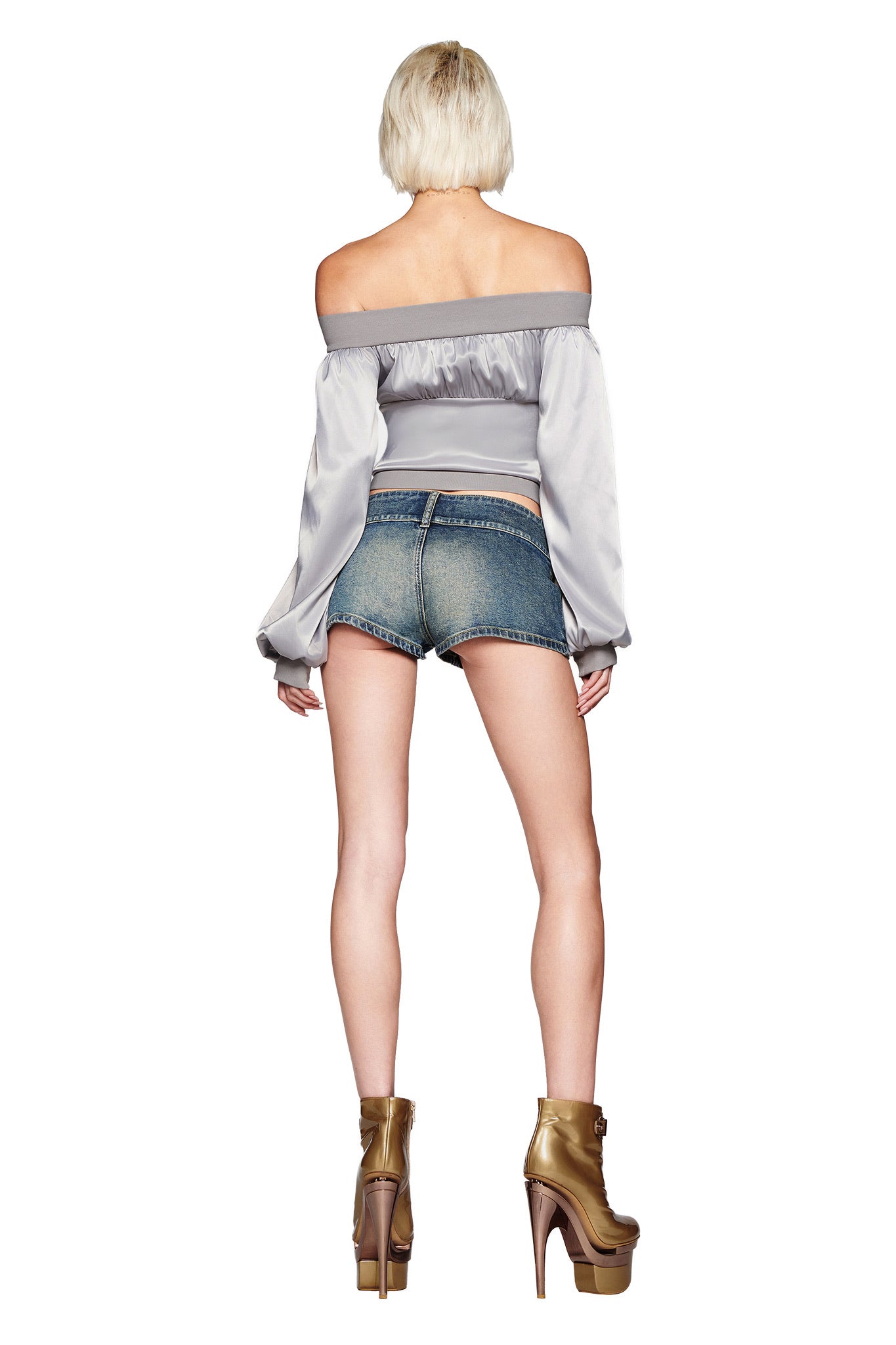 A person with short blonde hair faces away, wearing the I.AM.GIA JORDANA TOP in silver—an off-the-shoulder style with oversized bell sleeves—paired with denim shorts and gold platform heels against a white background.