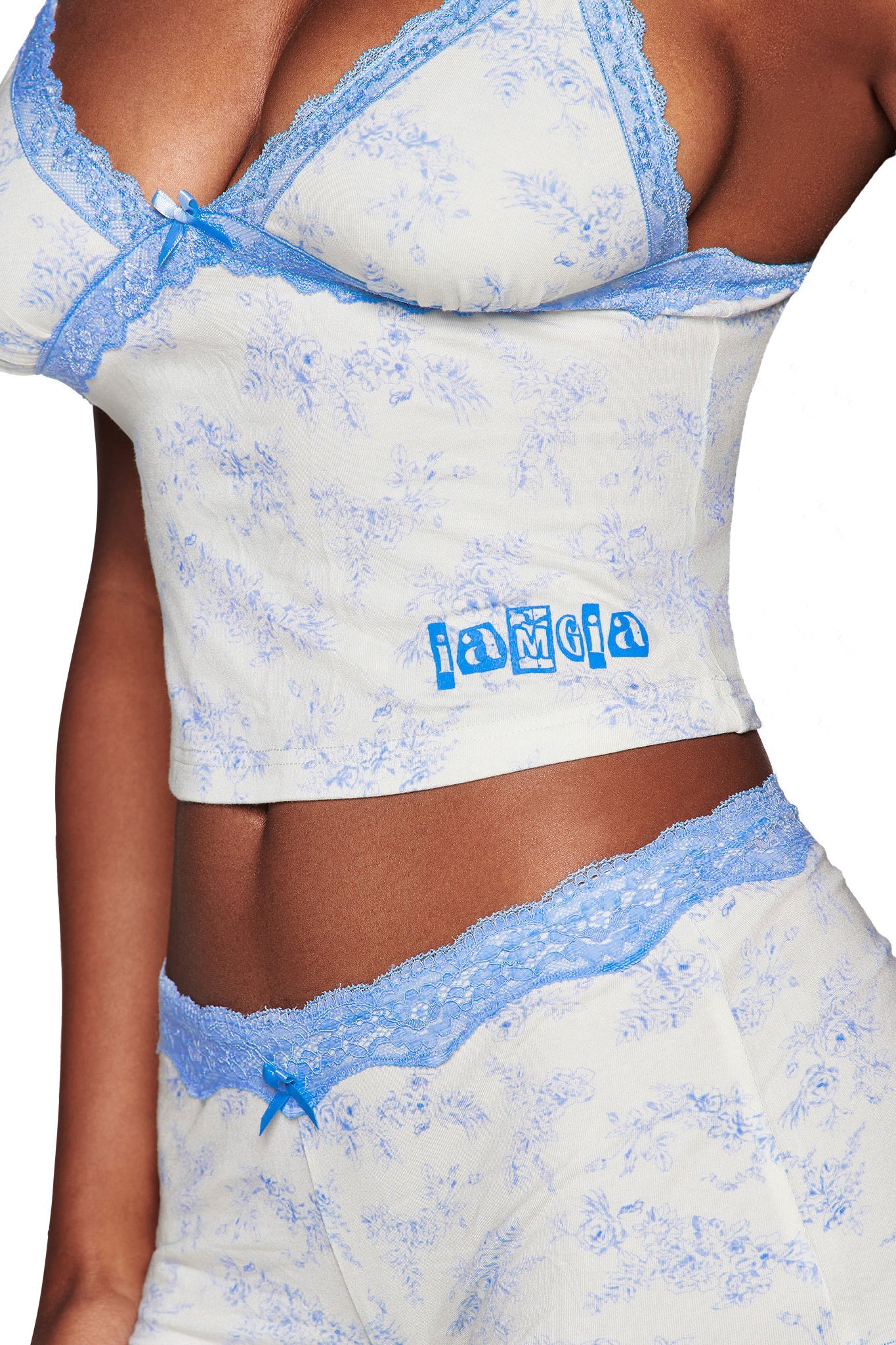 A person wears the I.AM.GIA CASSIA SHORT - BLUE TOILE set, featuring a light-colored toile floral print with blue lace trim and "JAMCIA" in blue on the white camisole.