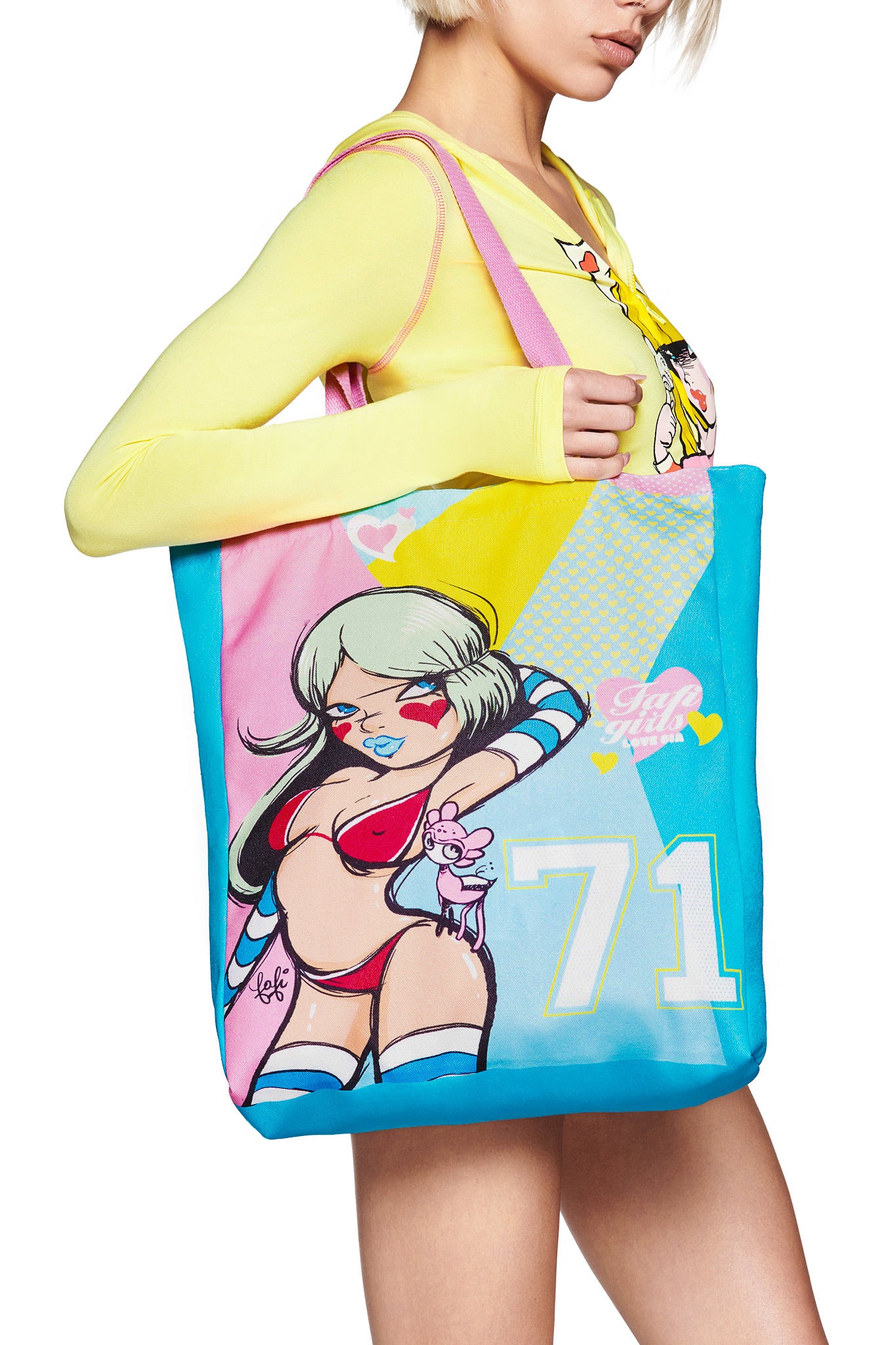 A person with short blonde hair carries the I.AM.GIA FAFI TOTE BAG - MULTI, decorated with Fafi artwork of a cartoon woman, hearts, and the number 71 in pastel colors. They pair it with a yellow graphic long-sleeve top.