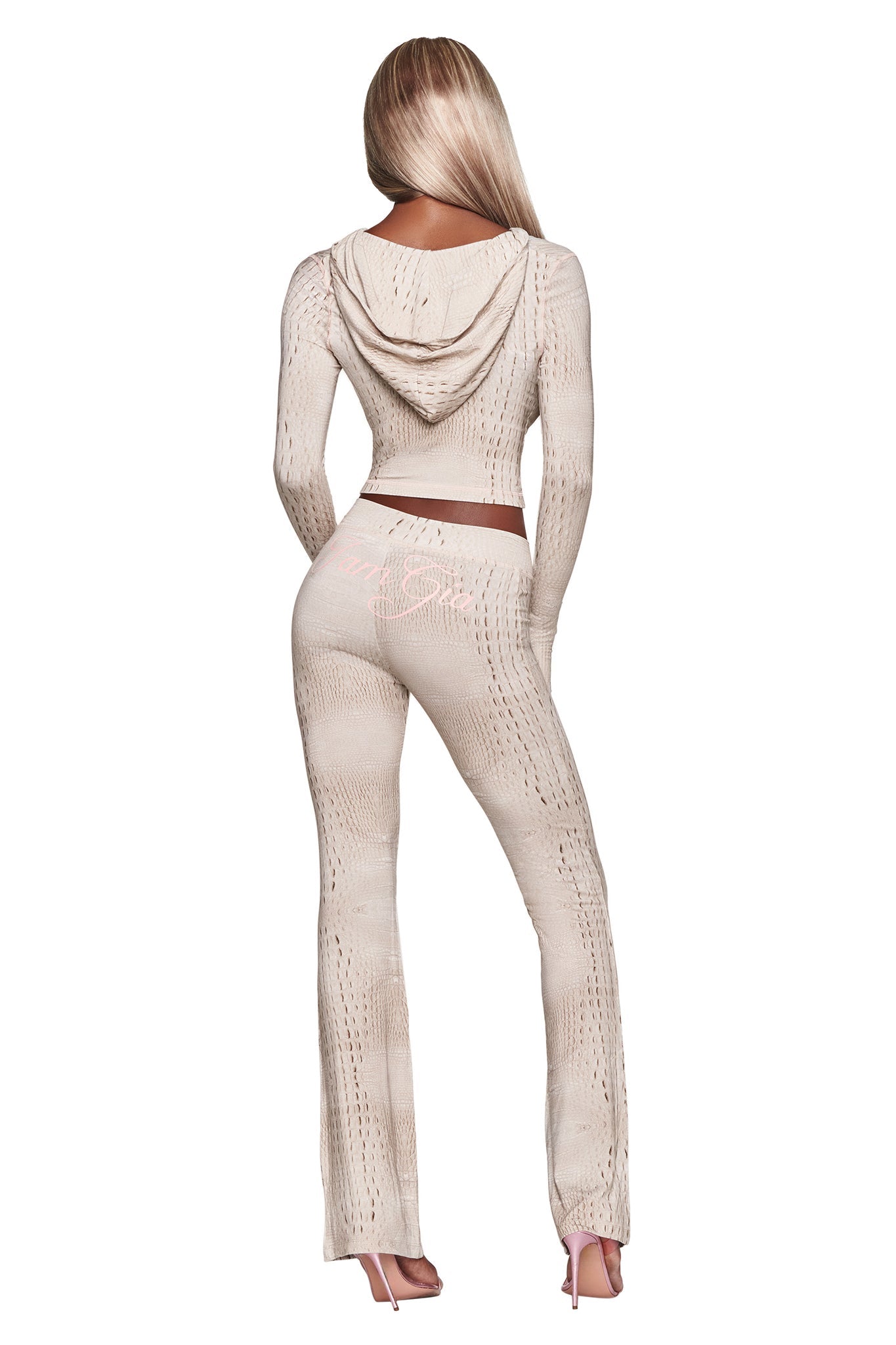 A woman with long blonde hair, standing with her back to the camera on a white background, wears a beige hooded crop top and matching I.AM.GIA BLARE TRACKPANT - HOT CROC flared pants paired with pink high heels.
