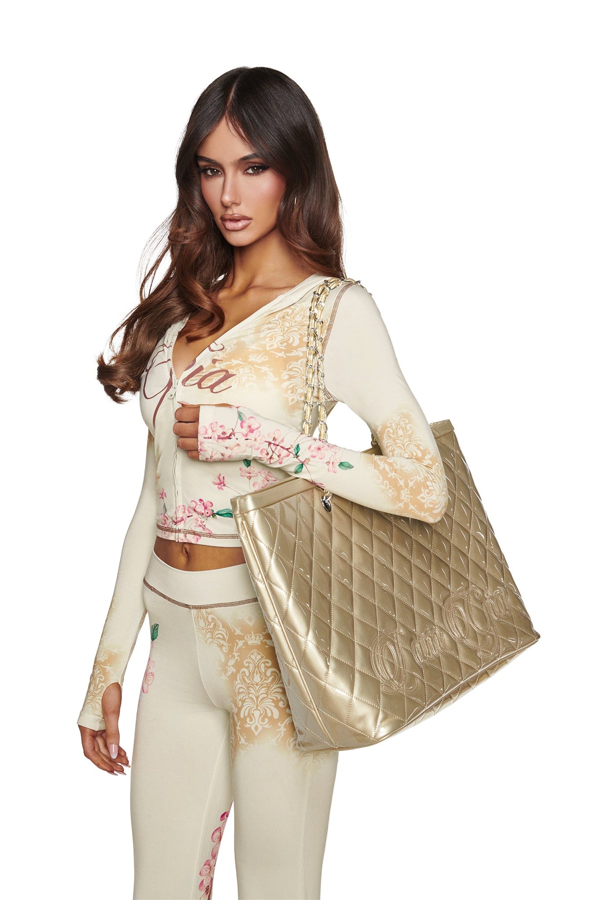 A woman with long brown hair wears a fitted, patterned cream outfit and carries the I.AM.GIA DANNI TOTE BAG in gold, featuring metallic finish and chain link straps, over her shoulder against a plain white background.