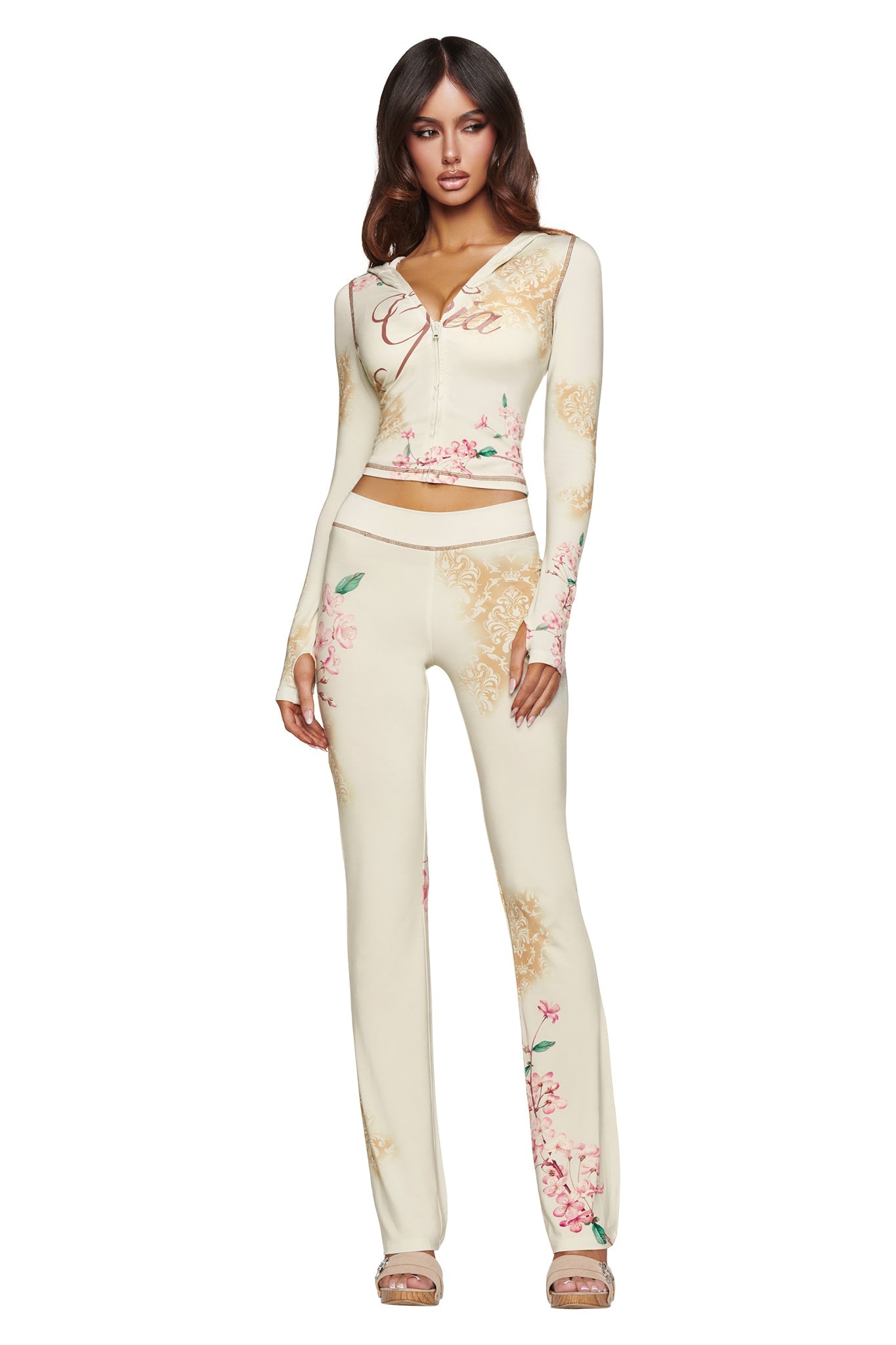 A woman stands against a white background wearing the I.AM.GIA BLARE TRACKPANT - BLOSSOM, a cream-colored, floral print two-piece set featuring a zip-up, long-sleeve crop top and high-waisted flare pants. She has long, wavy brown hair and is wearing sandals.