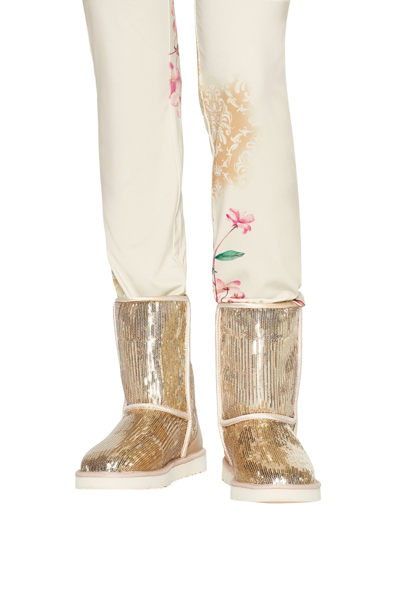 A person wearing cream-colored pants with floral patterns and the ELVIRA SEQUIN BOOT in Gold by I.AM.GIA, featuring striking gold sequins and a cozy faux fur lining, stands on a white background.