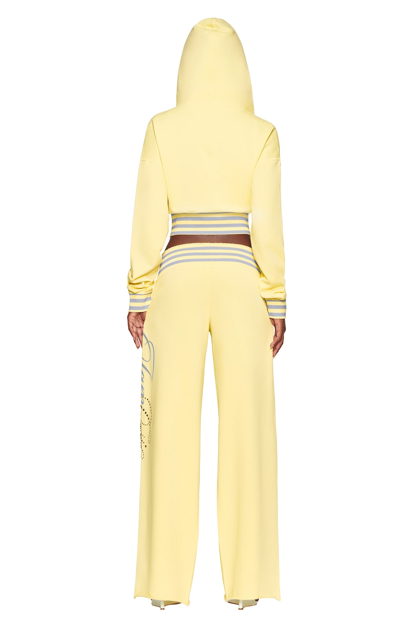 A person wearing the I.AM.GIA VEDA PANT - YELLOW, a wide-leg silhouette with a striped waistband and lettering on the left pant leg, paired with a matching yellow hooded sweatshirt, is standing with their back to the camera.