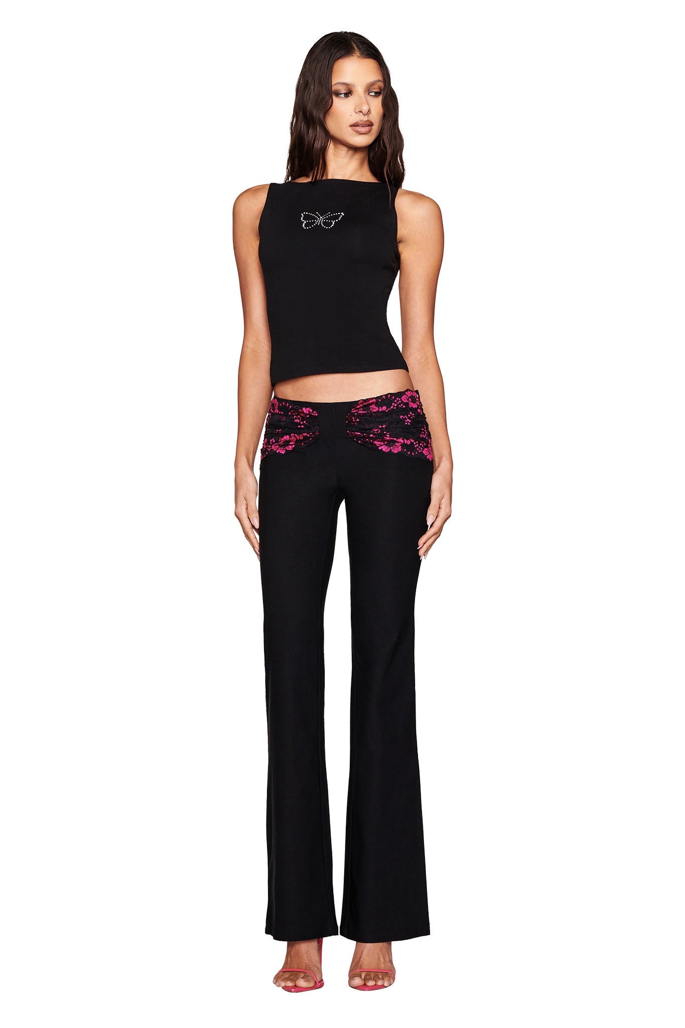 A woman models I.AM.GIA's sleeveless black top with a butterfly design and ANGELINA PANT - BLACK : LACE, featuring magenta lace panels. She stands with arms at her sides in pink heels against a white background.