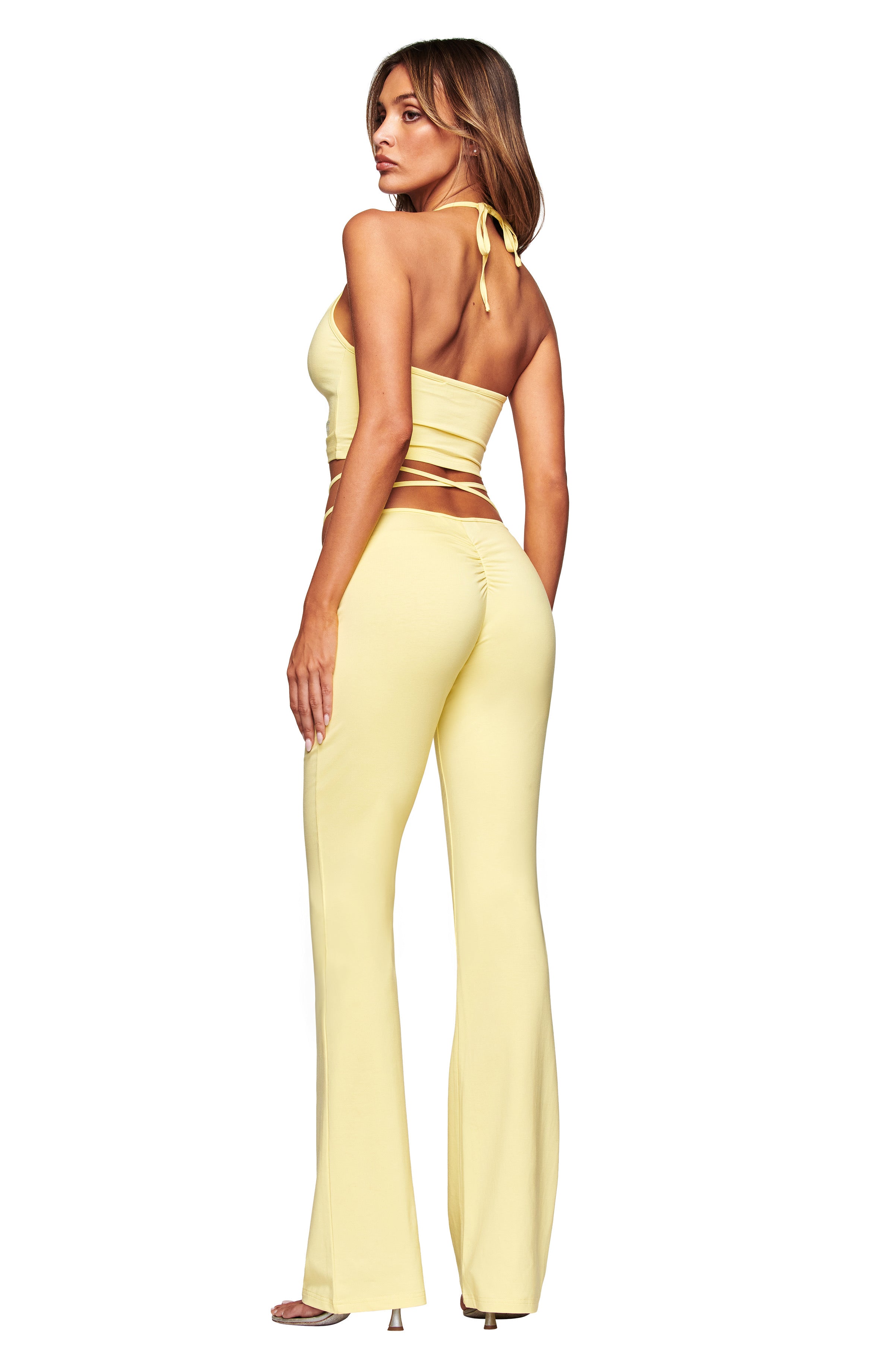 A woman stands facing away, wearing the I.AM.GIA HALO TOP in butter yellow with back ties and matching high-waisted flared pants, looking over her shoulder with long hair down and high-heeled sandals, set against a white background.