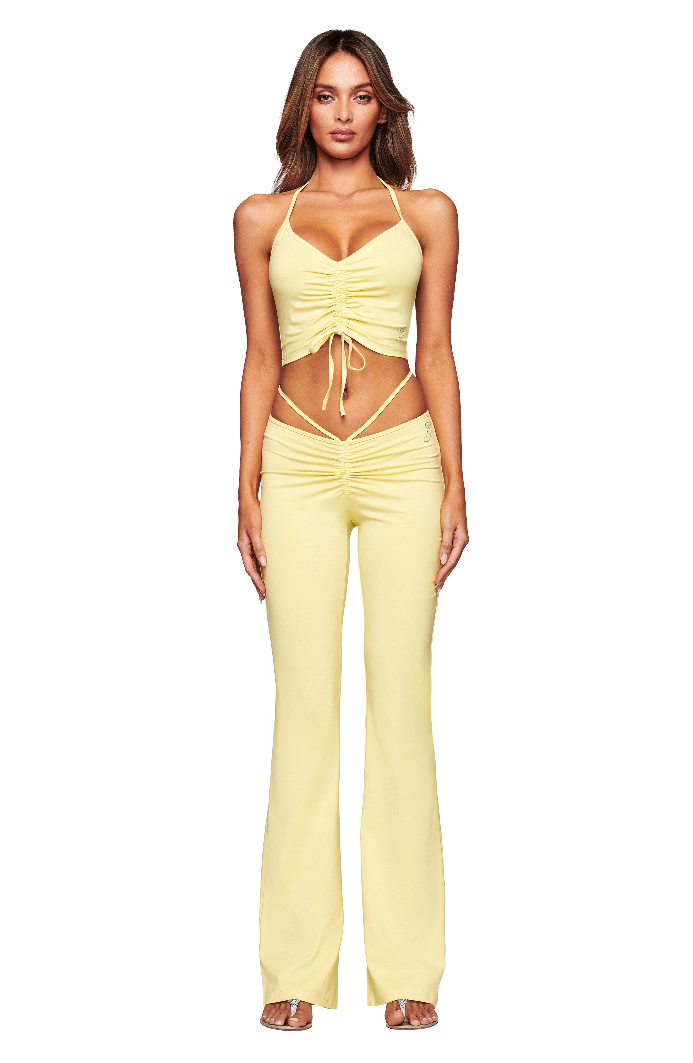 A woman poses confidently against a plain white background, wearing the I.AM.GIA HALO TOP in butter yellow—a halter style with ruched detailing—paired with matching high-waisted, flared pants. She has long, wavy hair and faces forward.