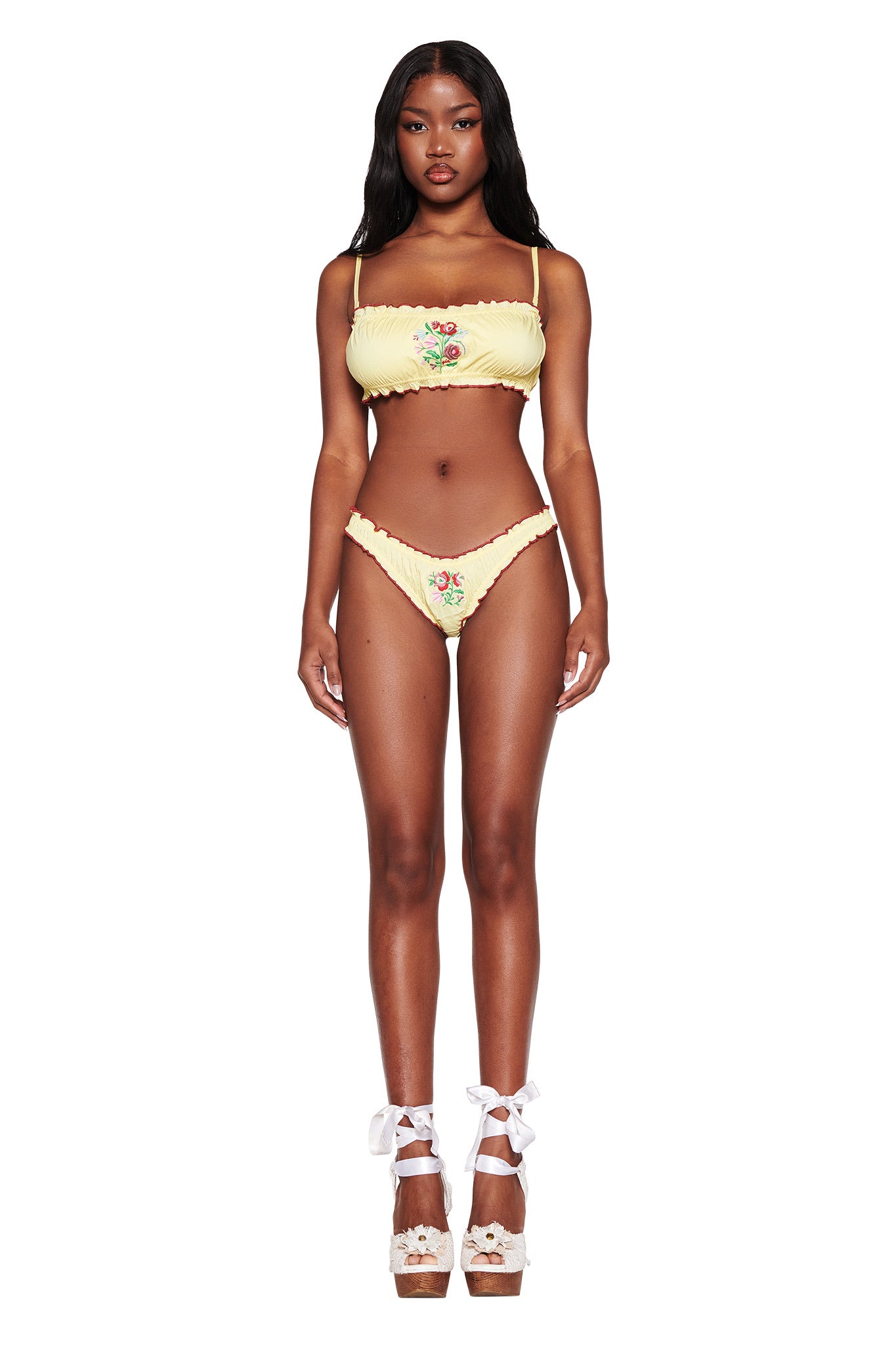 A model poses in the I.AM.GIA CLOVER SET - YELLOW, highlighting the bandeau top and high-waisted bottoms with floral embroidery. She complements the ensemble with ribbon-tie platform heels against a plain white background.