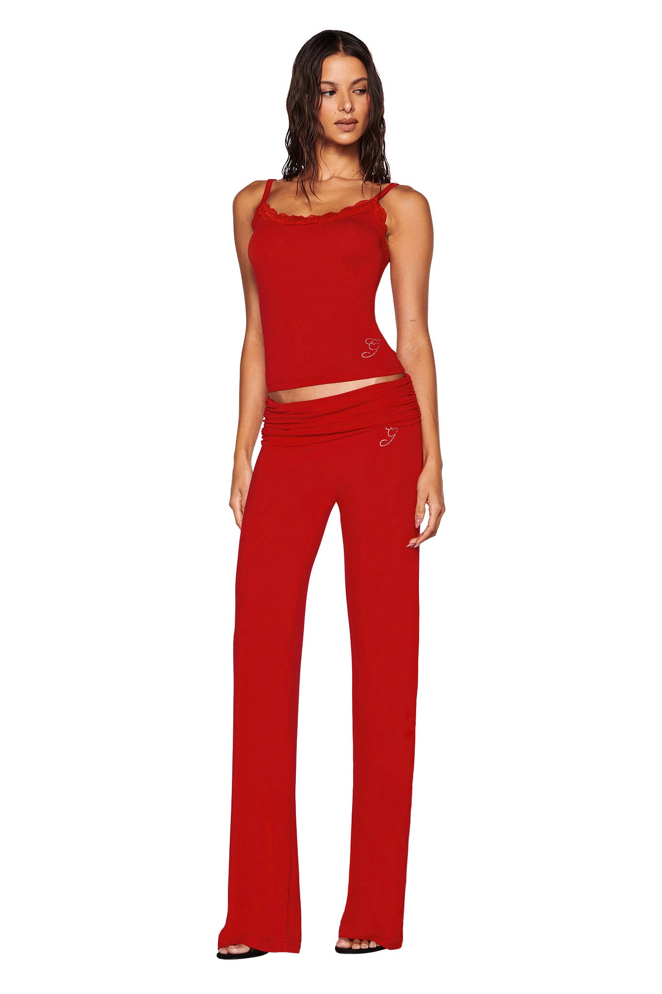 A woman is dressed in a red sleeveless ERIN TOP from I.AM.GIA, perfectly paired with matching red pants. One hand rests on her hip while the other hangs relaxed at her side. Her long hair cascades as she looks toward the camera, and subtle heart designs enhance the charm of this bamboo fabric ensemble.