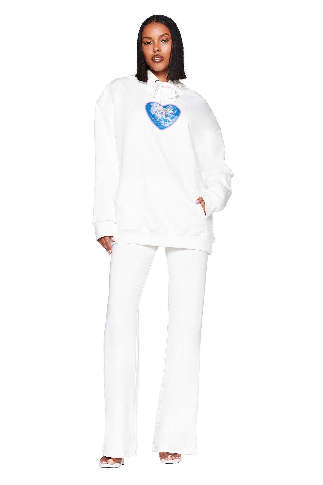 Clad in I.AM.GIA's Harmony Hoodie - White with a blue heart logo and matching pants, an individual with straight, shoulder-length hair stands against a plain white backdrop, accessorized with open-toed heels, showcasing chic style.