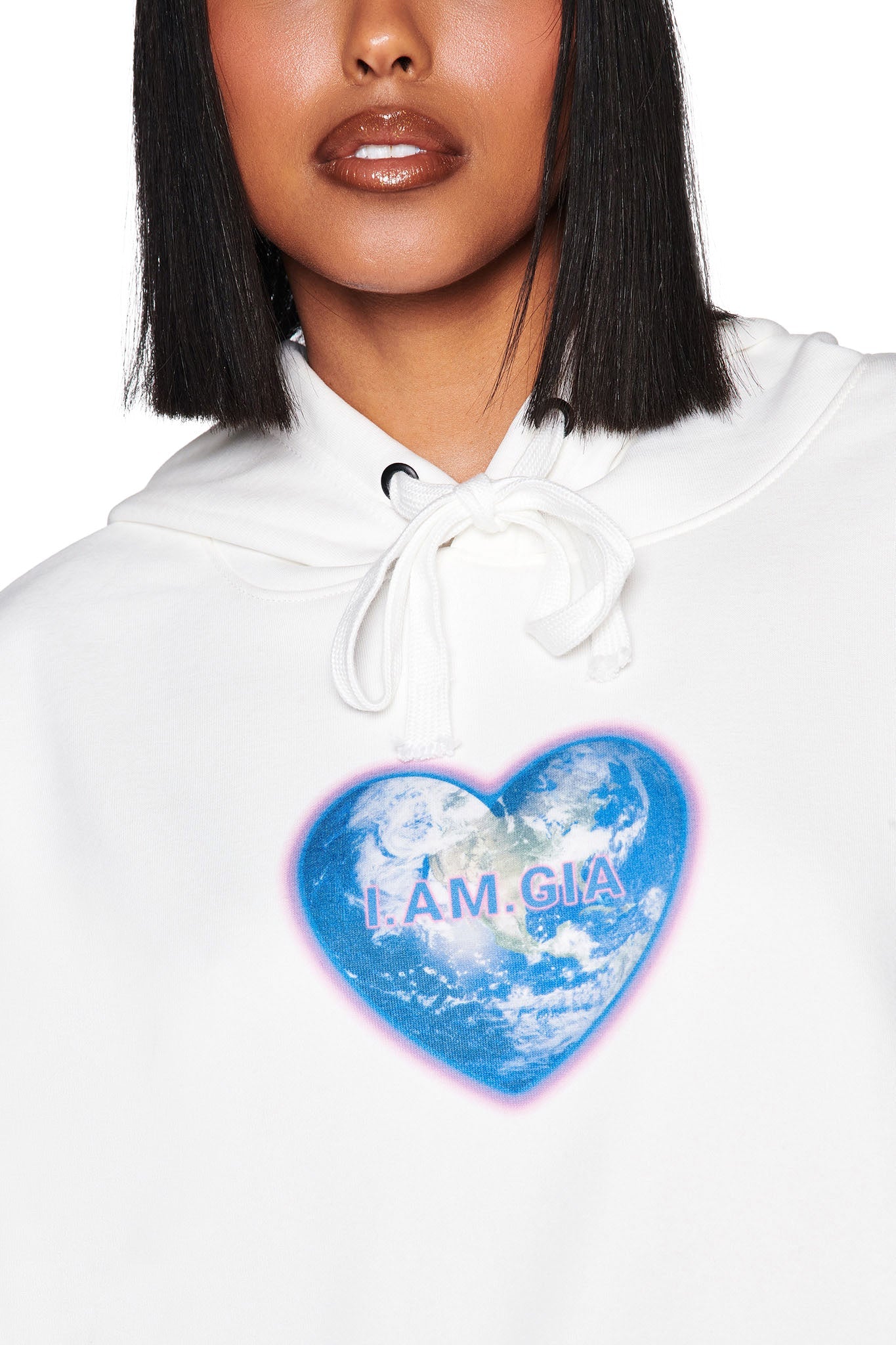 A person is styled in the I.AM.GIA Harmony Hoodie in white, featuring a heart-shaped Earth graphic with "I.AM.GIA." text. Their shoulder-length straight hair and glossed lips enhance their look against a minimalist background, highlighting the chic sweatshirt.