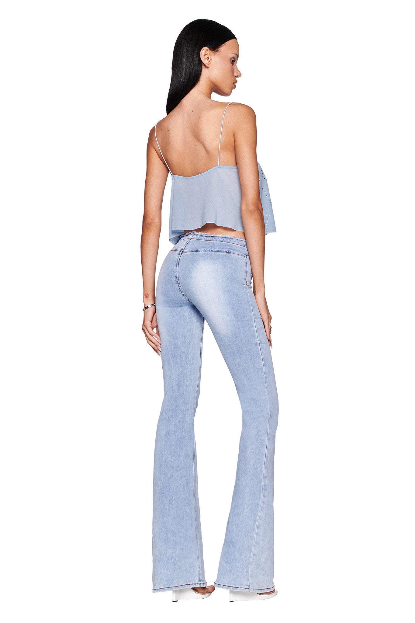 A woman with long dark hair, wearing the I.AM.GIA MINA TOP in baby blue and high-waisted light-wash flared jeans, stands against a plain white background. She is turned slightly to the side in clear high heels.