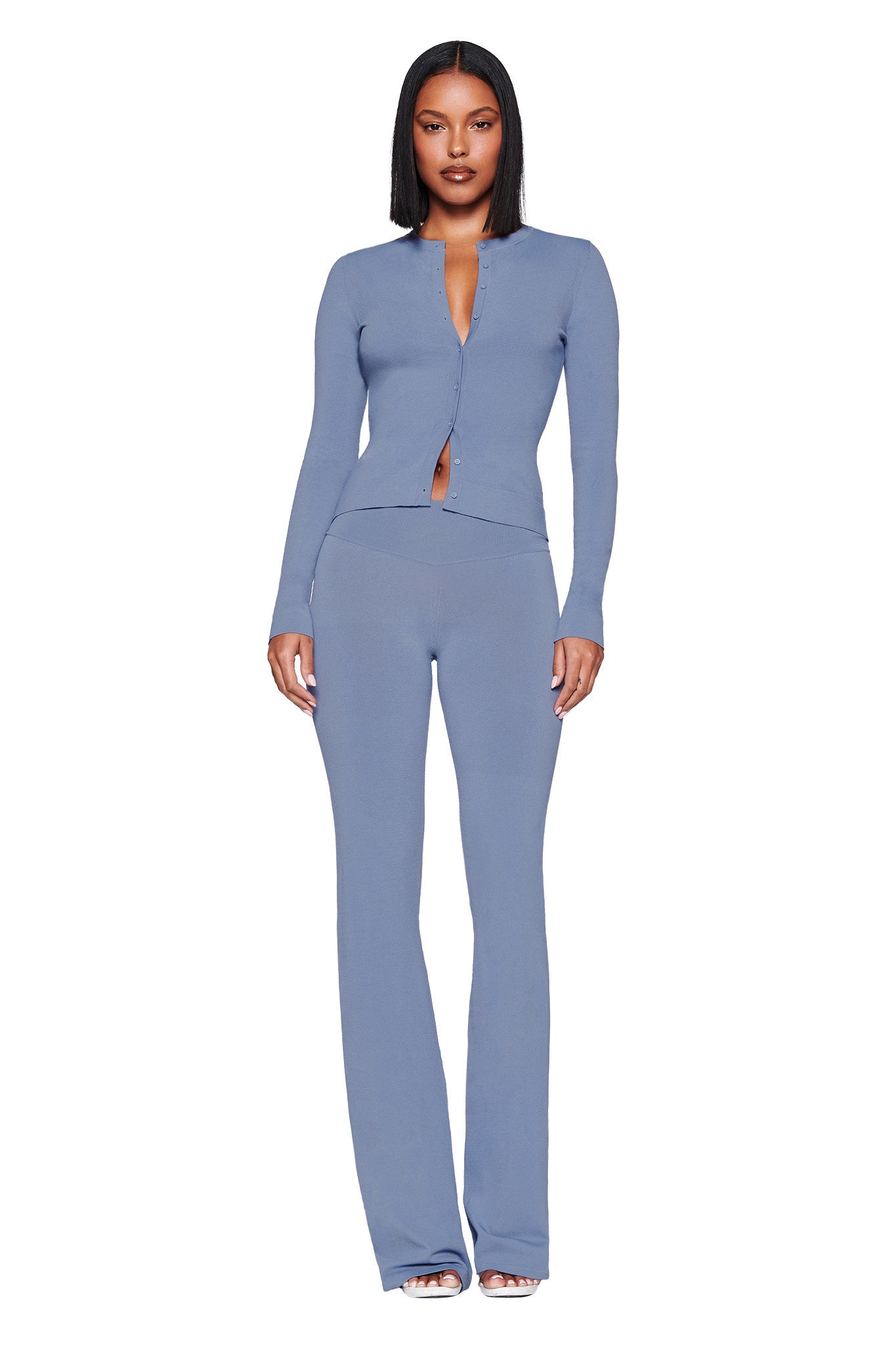 A person with shoulder-length black hair poses confidently against a white background, wearing a fitted long-sleeve top and I.AM.GIA's Audrina Pant in Steel Blue. The knit outfit features a V-neck and an open waist segment.