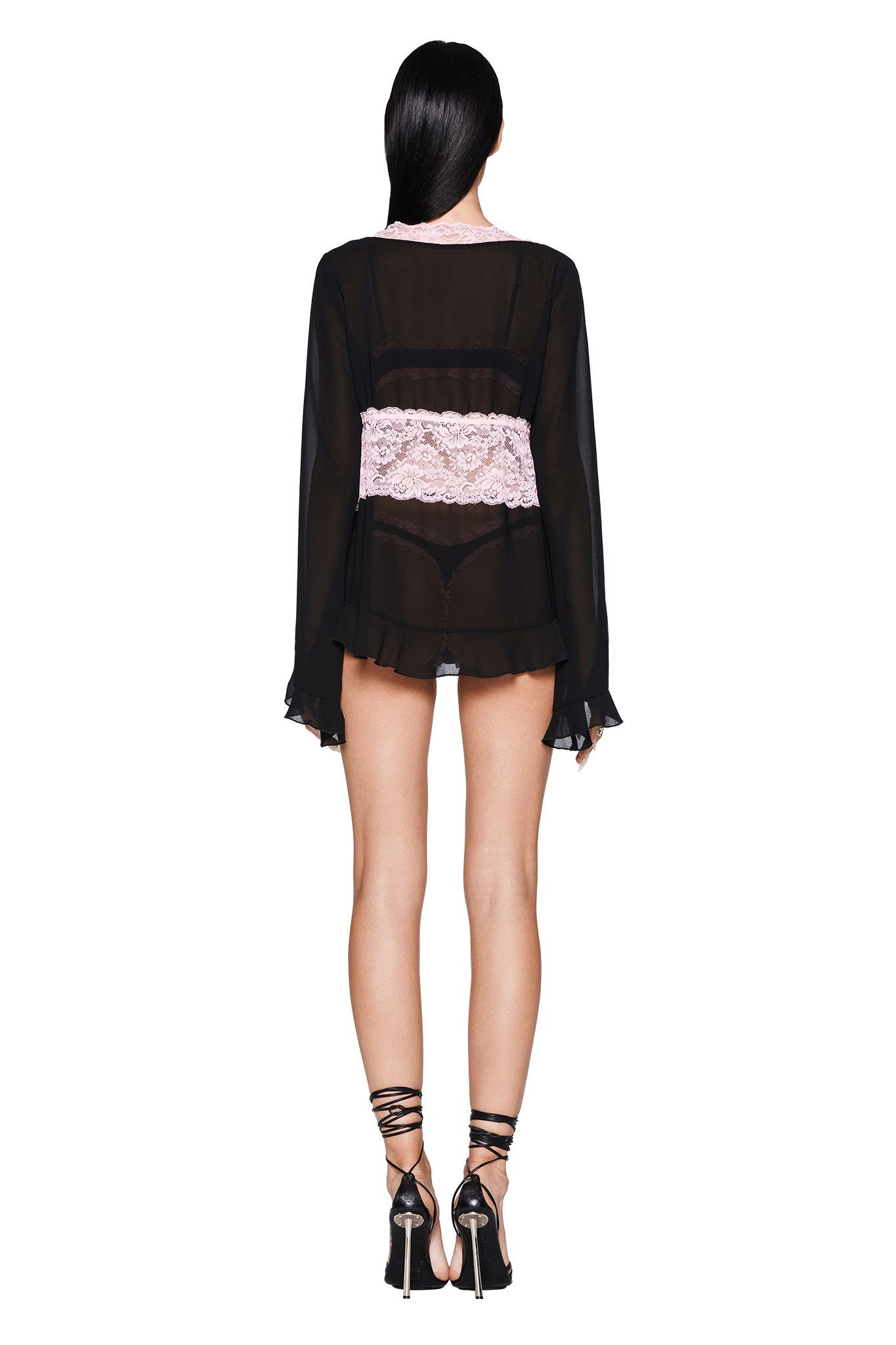 A rear view showcases a person with long black hair wearing the I.AM.GIA JUNIPER ROBE THREE-PIECE SET in black, featuring pink lace details around the waist for an elegant touch. They are paired with black high-heeled sandals that have ankle straps, all set against a plain white background.