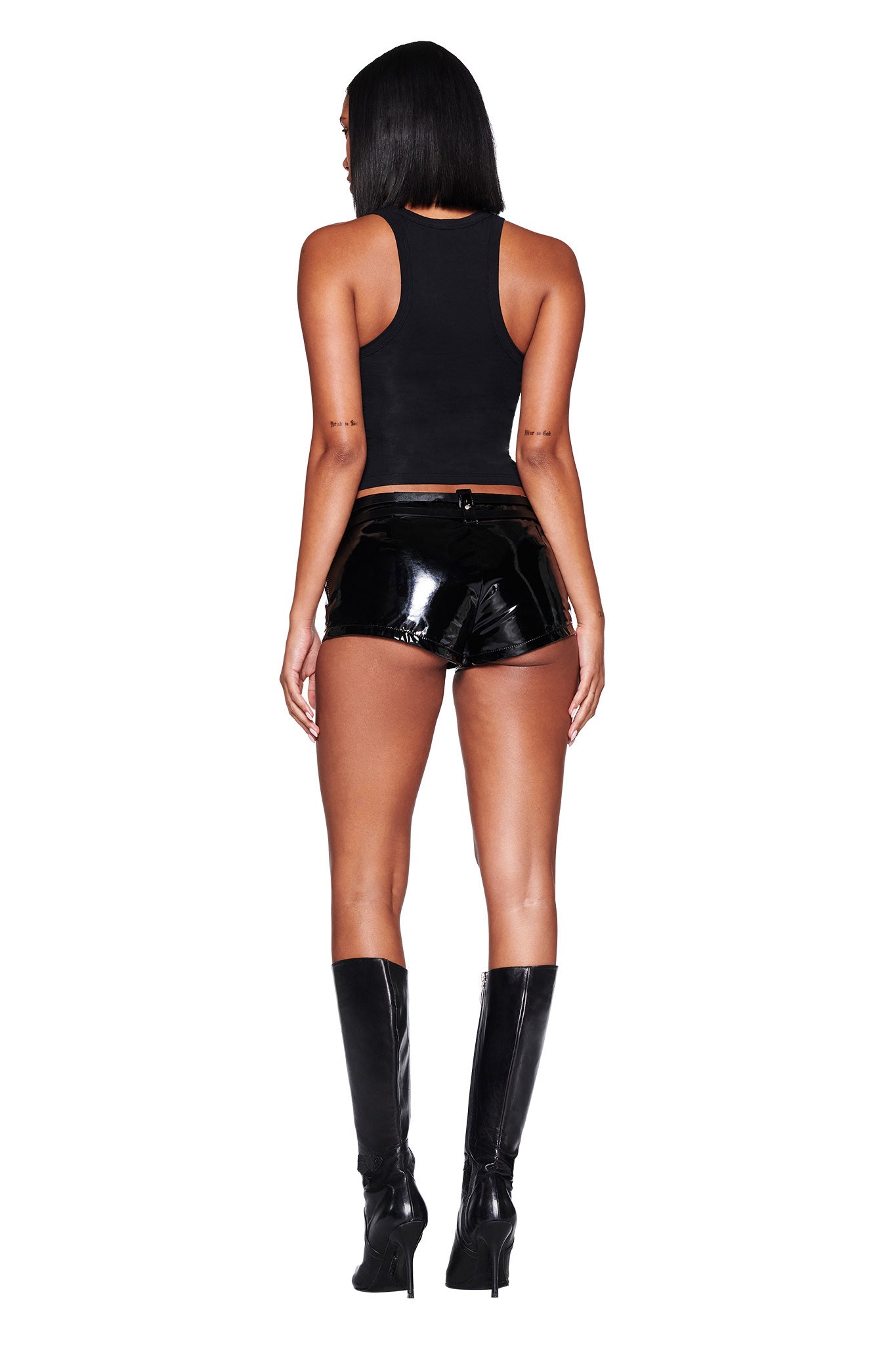 A person with long, straight hair stands facing away in a bodycon fit I.AM.GIA CAPRICORN ZODIAC TANK - BLACK, shiny black shorts, and knee-high black boots. The sleek look contrasts with the plain white background.
