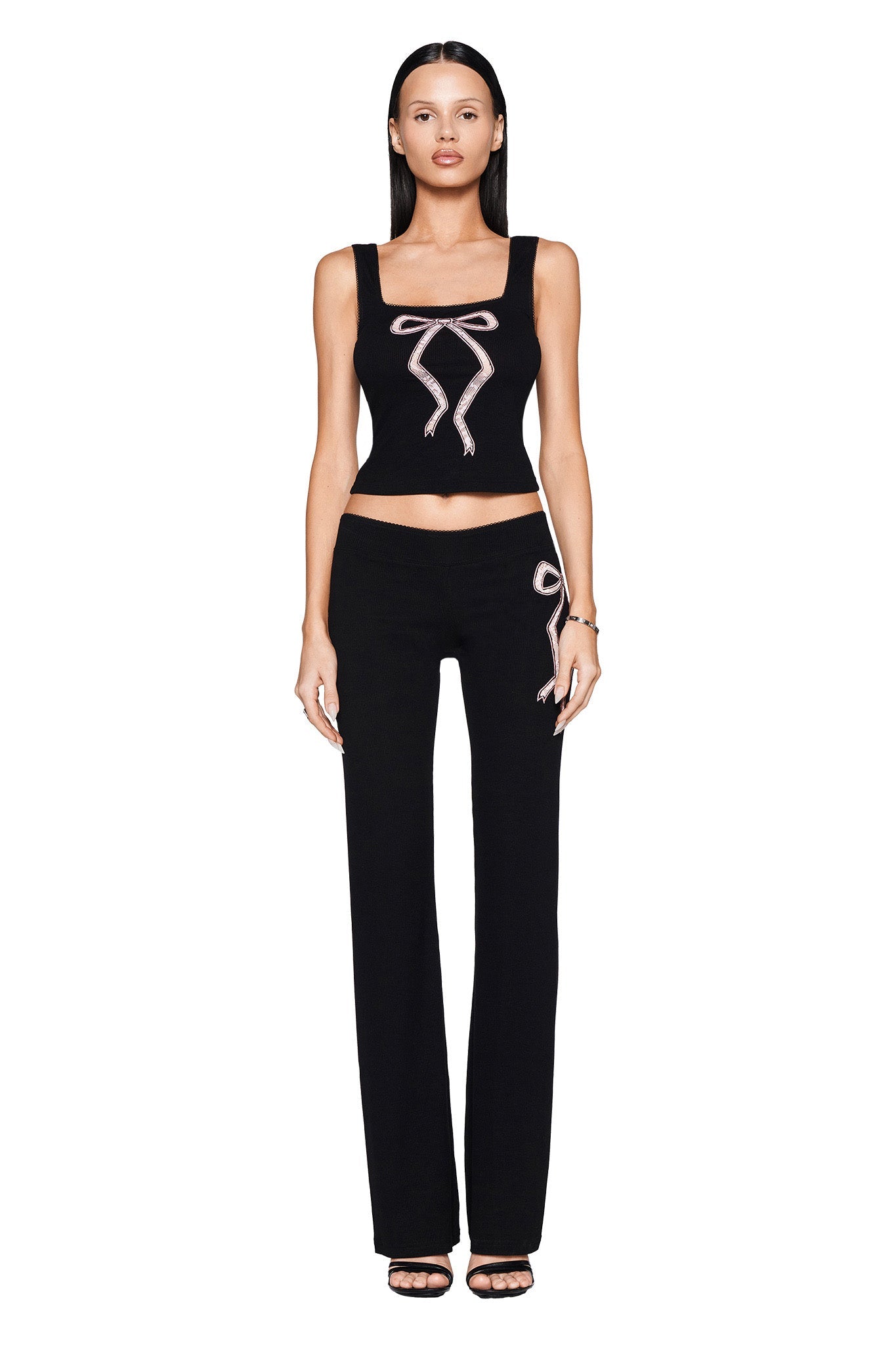 A woman stands facing the camera against a white background, wearing the ASTRID PANT in black from I.AM.GIA. This outfit, which pairs chic sleepwear vibes with streetwear fashion, showcases pink ribbon-like accents on the chest and hip area and features a subtle flared leg design. She completes her look with black sandals.