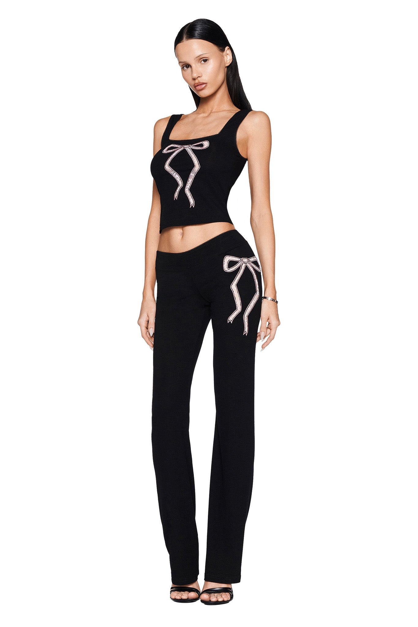 Wearing the ASTRID PANT - BLACK from I.AM.GIA, a woman poses gracefully in a chic black two-piece outfit, showcasing intricate ribbed knit fabric with decorative bow accents on the top and elegantly flared leg pants. Her long black hair cascades down as she stands confidently in black heels against a pristine white background.