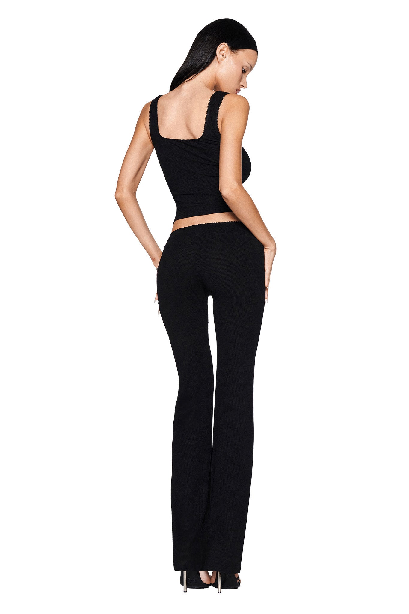 A person with long black hair stands facing away, showcasing the I.AM.GIA sleeveless ribbed knit top paired with the sleek ASTRID PANT in black and high heels. The minimalist design of the outfit is accentuated against a plain white background.