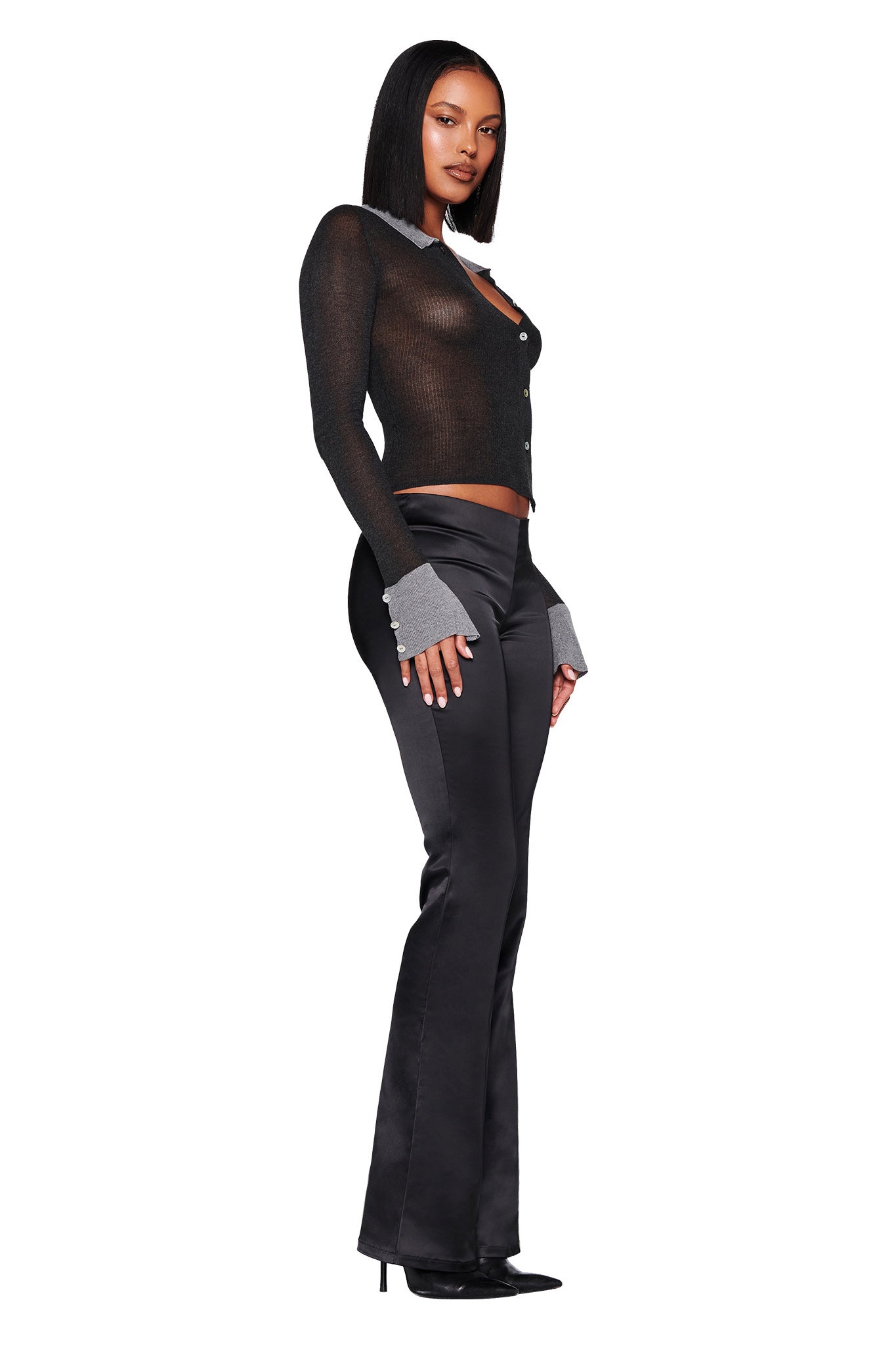 A confident individual poses against a white backdrop, clad in the stylish I.AM.GIA Davina Top in black with true knit fabrication, paired with black satin pants and pointed heels. The semi-transparent top boasts grey cuffs and pearl shell buttons for an added touch of sophistication.