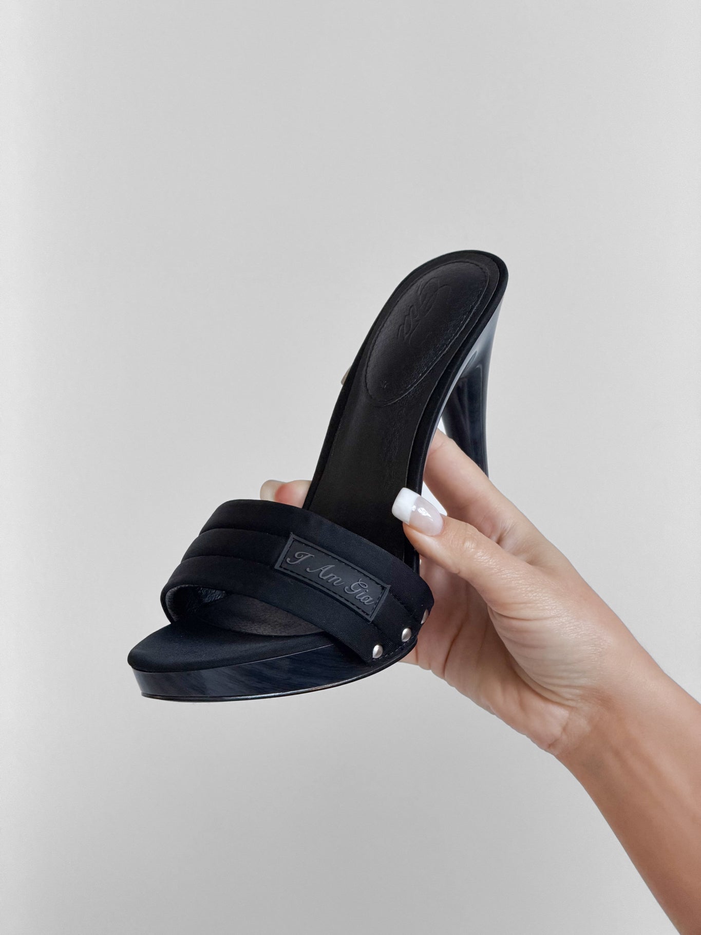 A hand with long, manicured nails holds the I.AM.GIA NICKY HEEL in black neoprene—a slip-on mule with an open toe, padded vamp strap, and sleek stiletto heel featuring a metal logo—set against a plain light gray background.