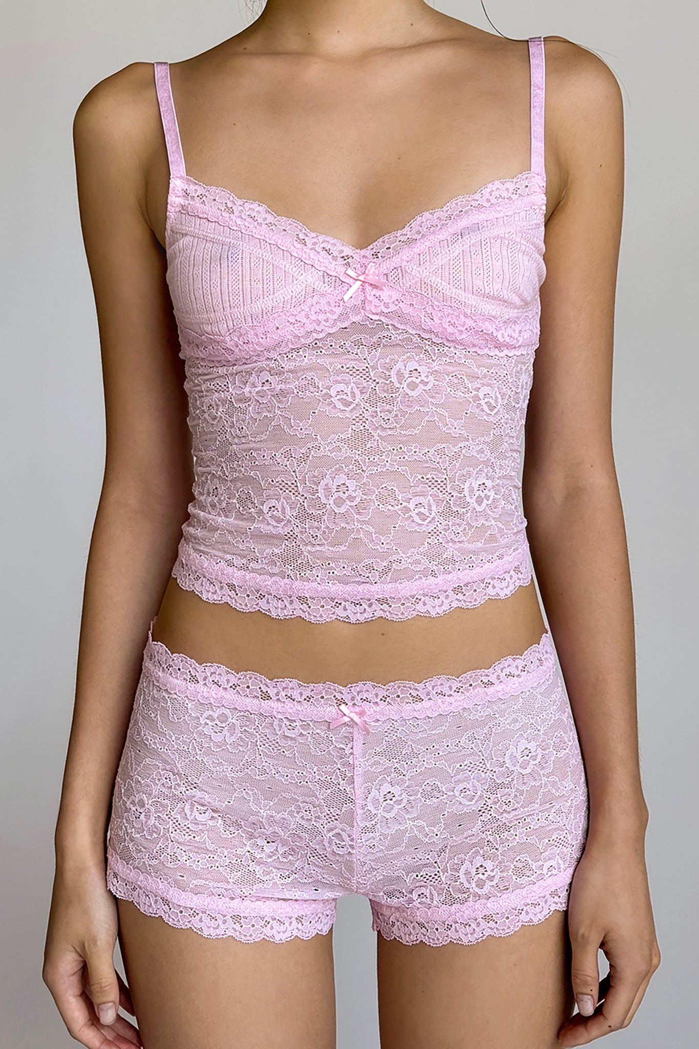 A person wearing the I.AM.GIA SERINA SHORT - PINK : LACE lingerie set, which includes a fitted camisole with delicate thin straps and lace detailing, paired with low-rise waist lace shorts that feature a small bow at the center of the waistband. The background is plain and neutral.