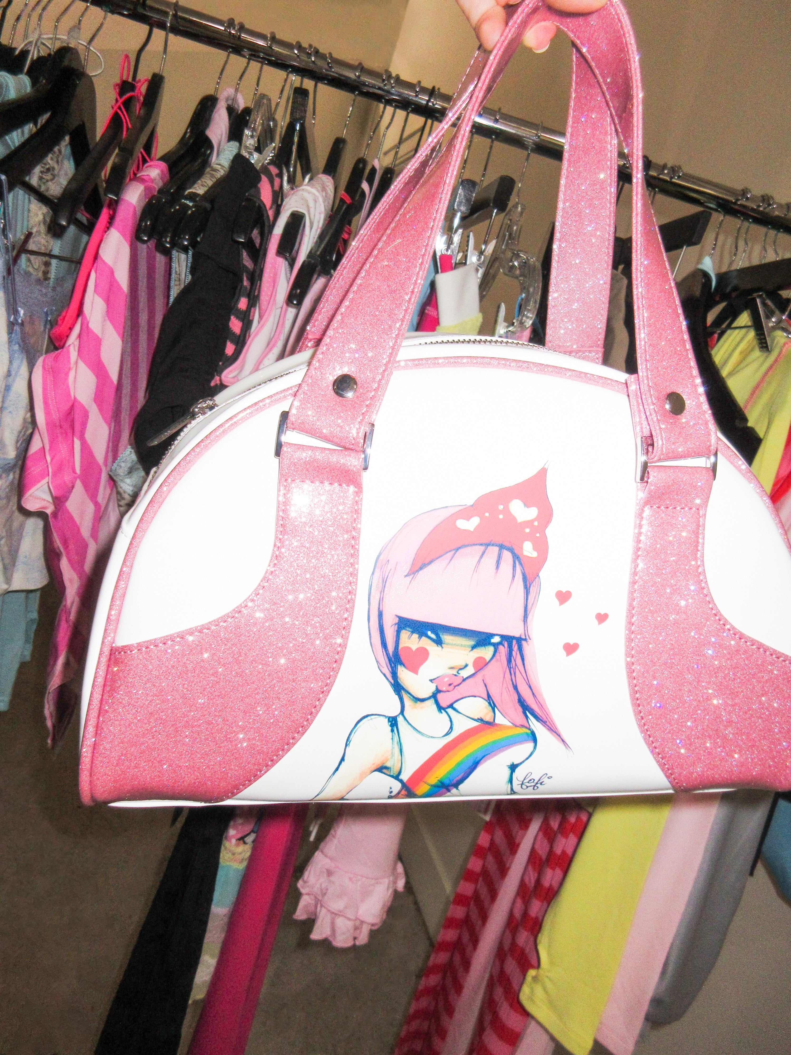 The FAFI BAG - PINK by I.AM.GIA is a sparkly vinyl bowling bag with pink glittery straps, featuring custom artwork of a pink-haired girl with face paint and a rainbow, displayed in front of colorful clothes on a rack.