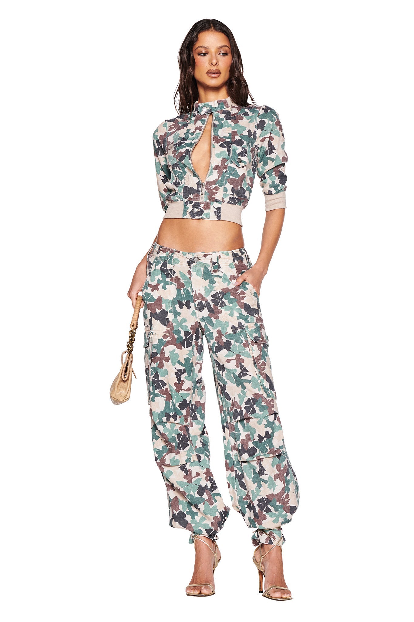 A woman poses confidently in I.AM.GIA's EFFIE CARGO PANT - FLUTTER CAMO. She is styled with a chic cropped top featuring a central cut-out and holds a beige purse while standing in high heels, her long hair cascading against a white backdrop.