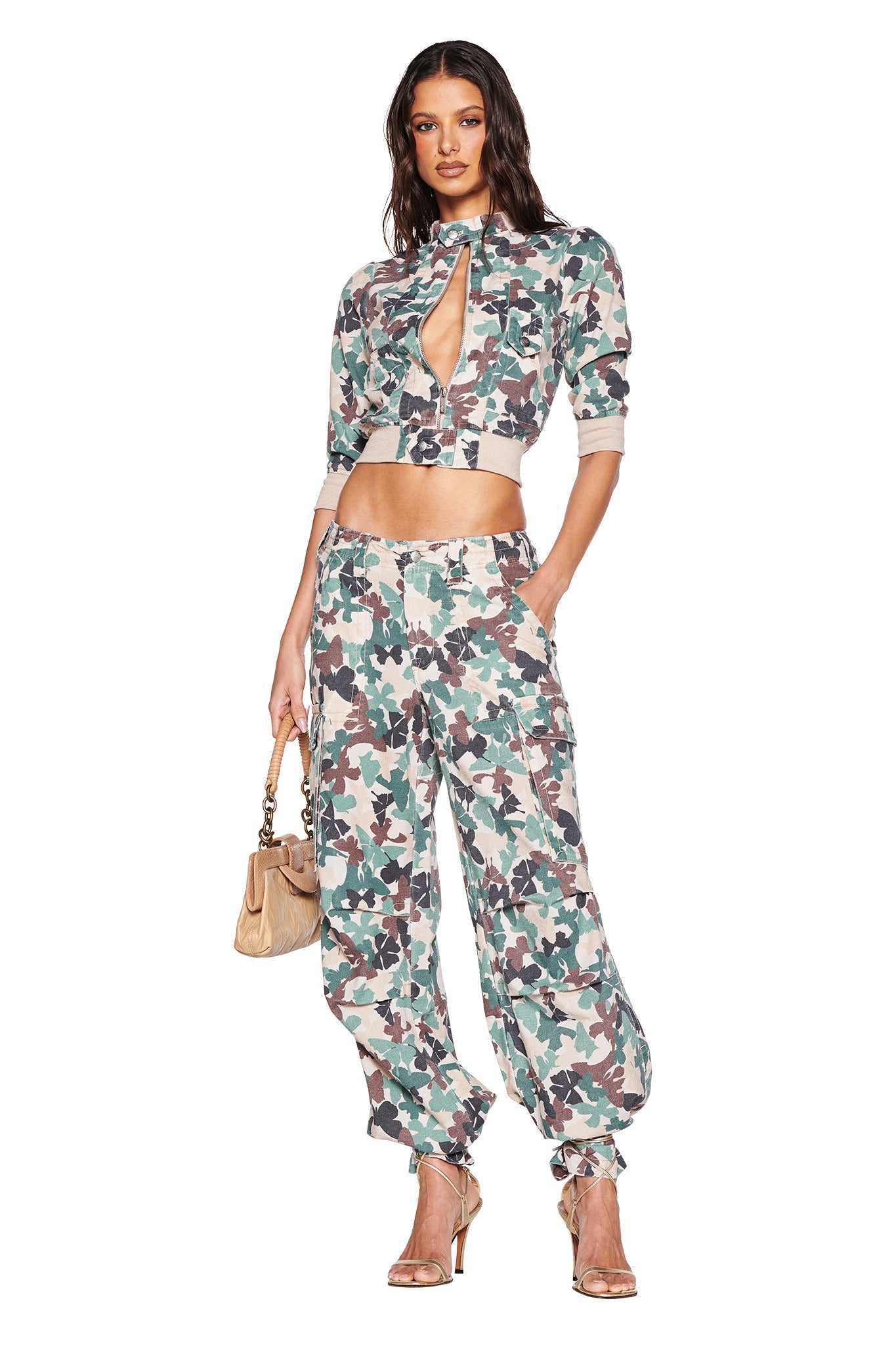 A person poses in an I.AM.GIA EFFIE JACKET - FLUTTER CAMO outfit featuring a butterfly camo print cropped top with a keyhole design and matching pants. They hold a beige handbag and wear gold high-heeled sandals against a white background.
