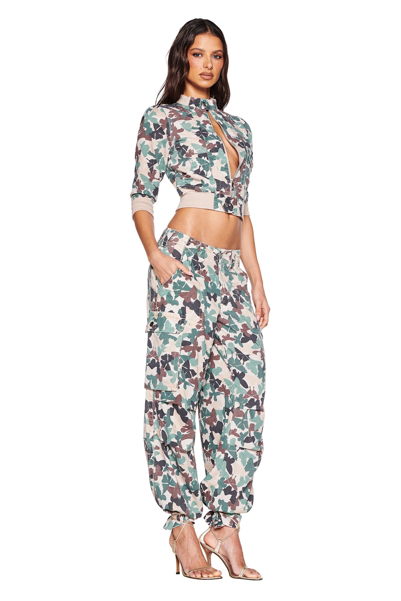 A woman strikes a pose in I.AM.GIA's EFFIE CARGO PANT - FLUTTER CAMO with a cropped jacket, complemented by stylish heels and her long hair cascading gracefully against a white background.