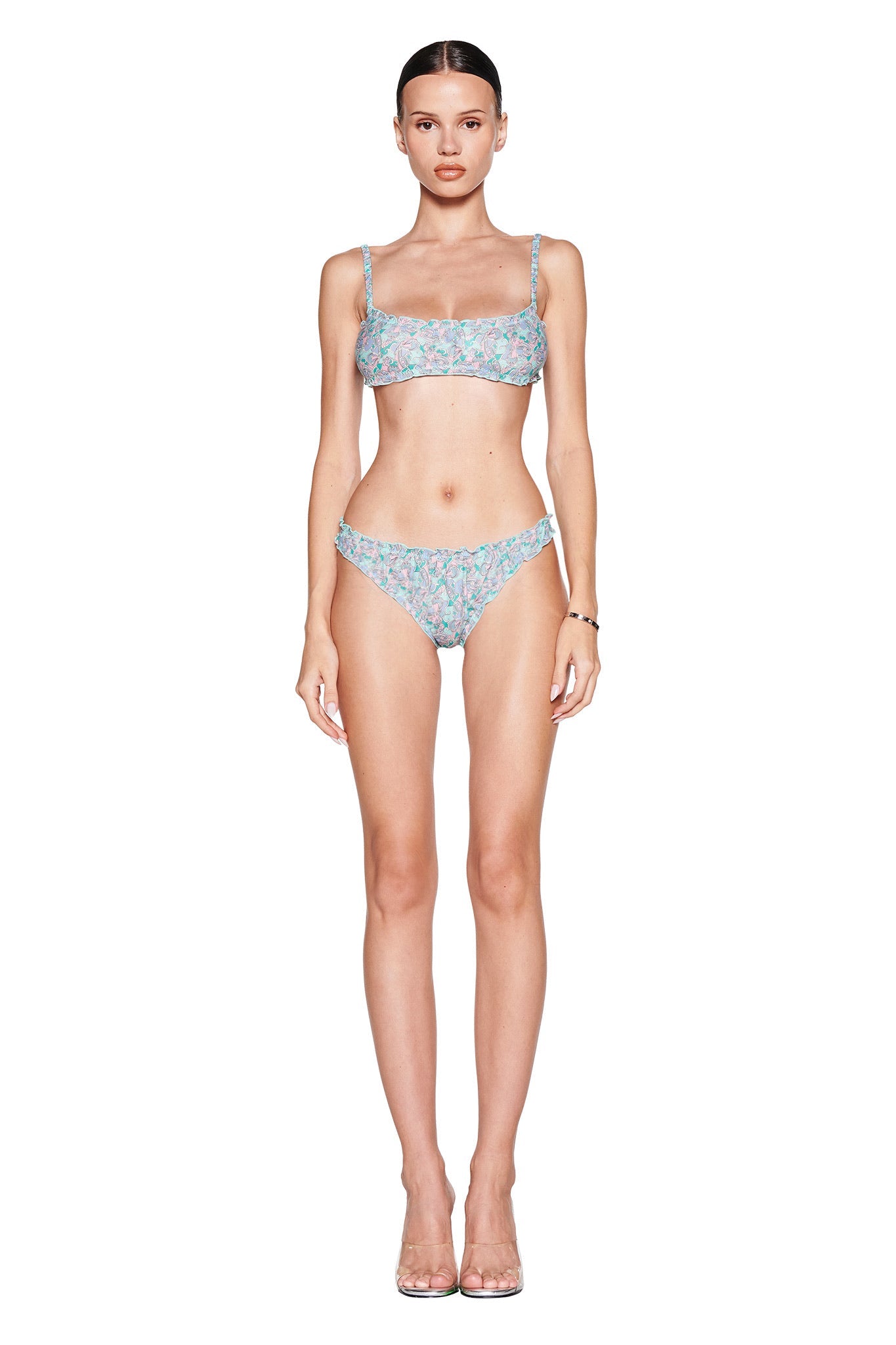 A person stands facing forward against a white background, wearing the I.AM.GIA JEAN Bralette Set adorned with a teal butterfly bloom pattern and ruffled straps. Transparent high heels complement the vintage-inspired style, and long, straight hair flows freely as arms relax by their sides.
