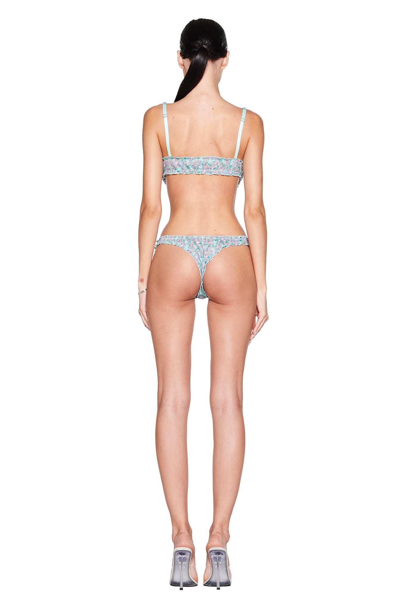 A person with long hair stands facing away, wearing the I.AM.GIA JEAN BRALETTE SET in Teal Butterfly Bloom, paired with clear high heels. The plain white background accentuates their graceful posture and vintage-inspired style.