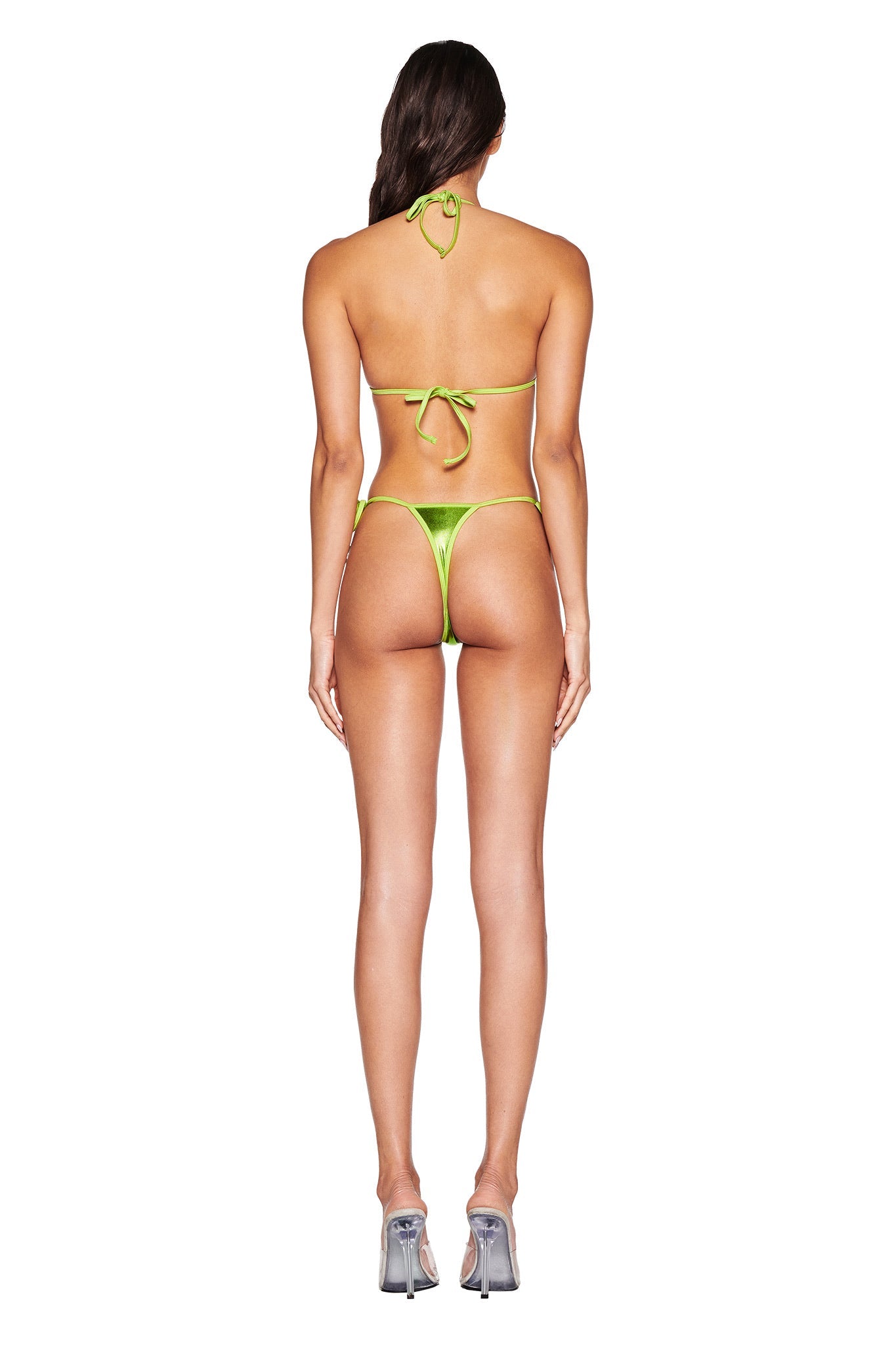 Rear view shows a person in an I.AM.GIA Alohi Bikini Bottom - Lime Green, paired with silver high heels. The bikini features a halter neck and back ties, crafted from metallic fabric with thong-style bottoms against a plain white background.