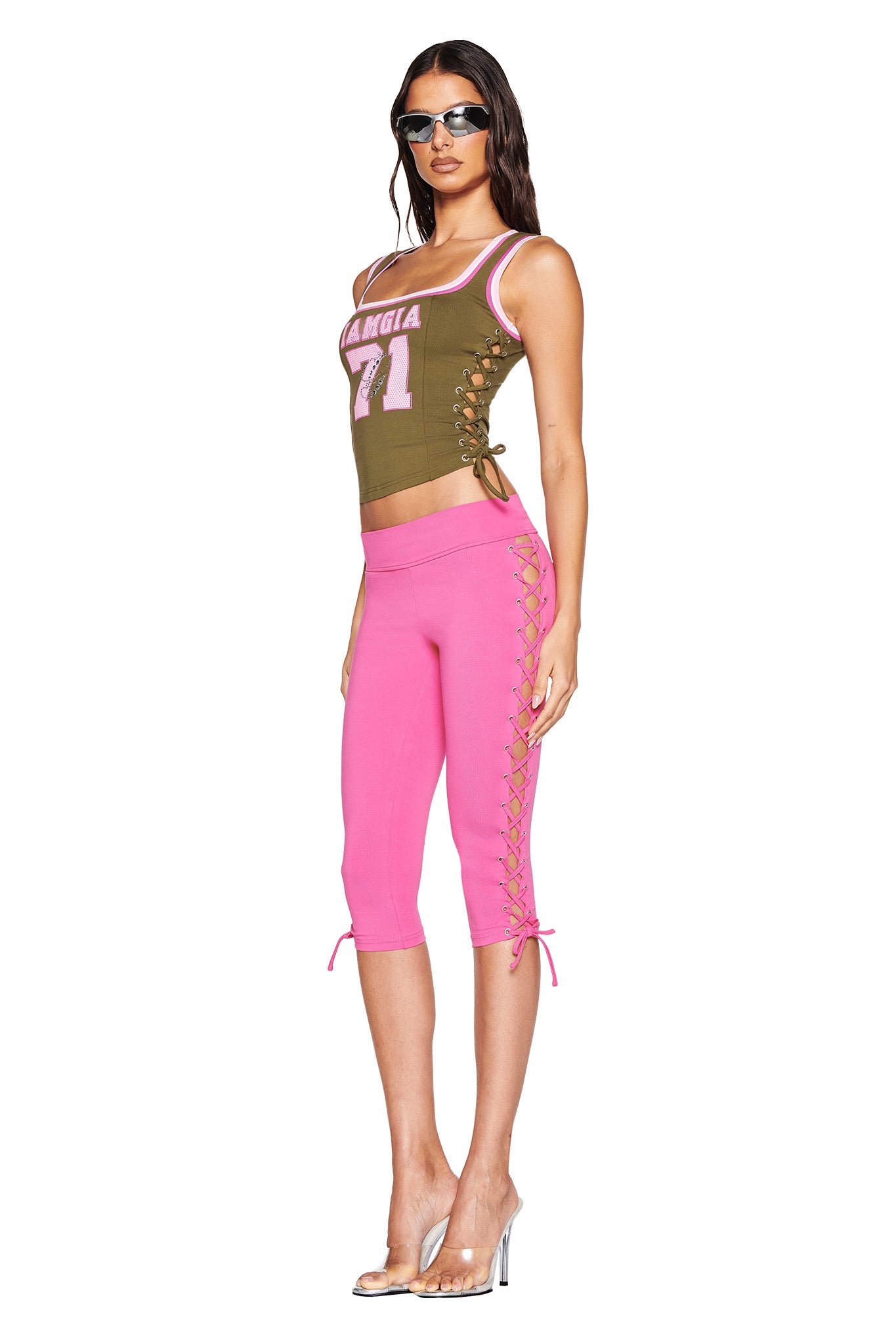 A woman wears an olive green top with "MANGIA 71" and lace-up details, paired with I.AM.GIA's TILA CAPRI in hot pink, accessorized with sunglasses and clear high heels. Her chic look pops against a pristine white backdrop, blending comfort and flair.