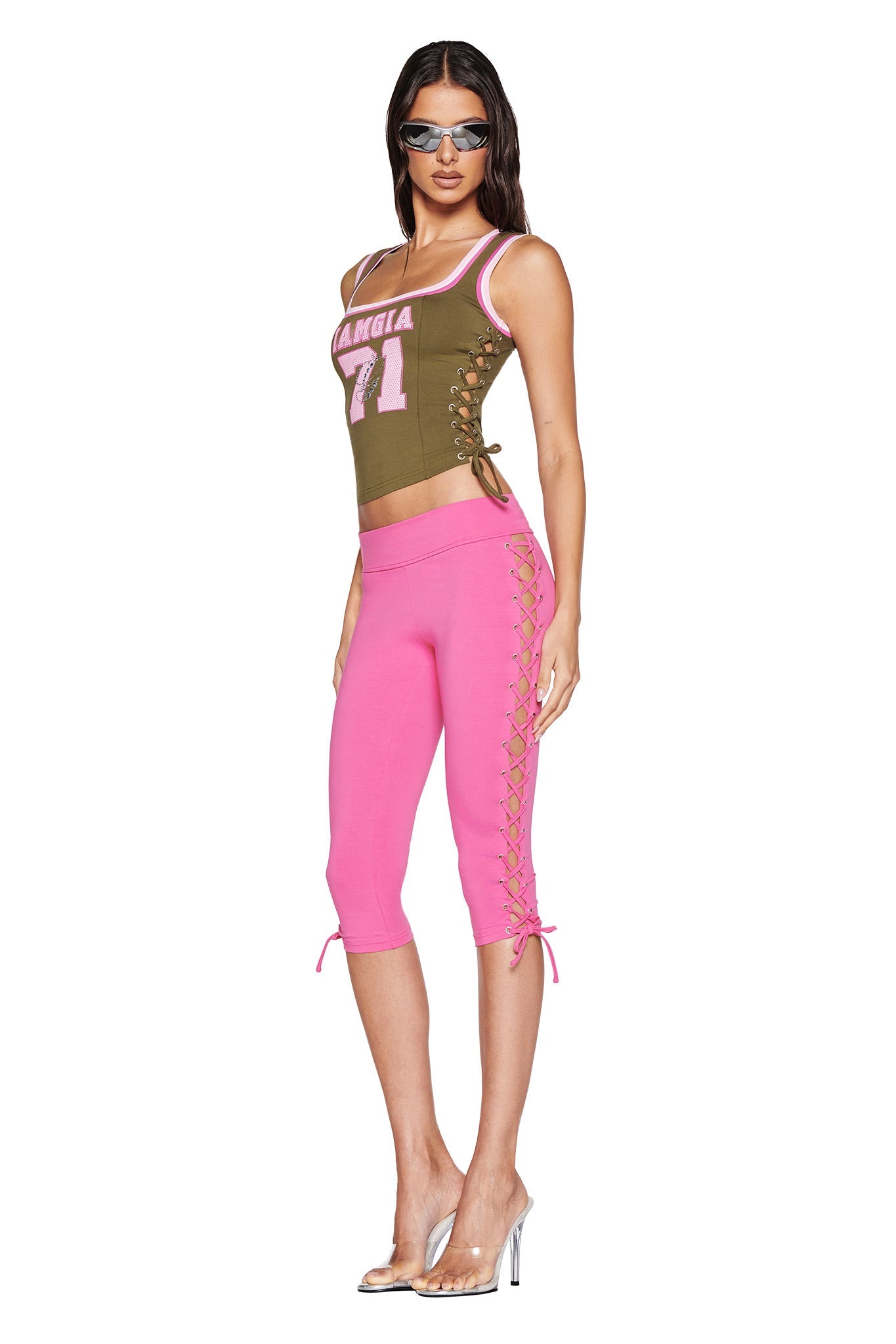 A woman confidently poses in high heels, wearing I.AM.GIA's olive green sleeveless top with "AMGIA 71" and the TILA CAPRI - HOT PINK pants. Her hair is down, accentuating the bodycon fit of her stylish ensemble.