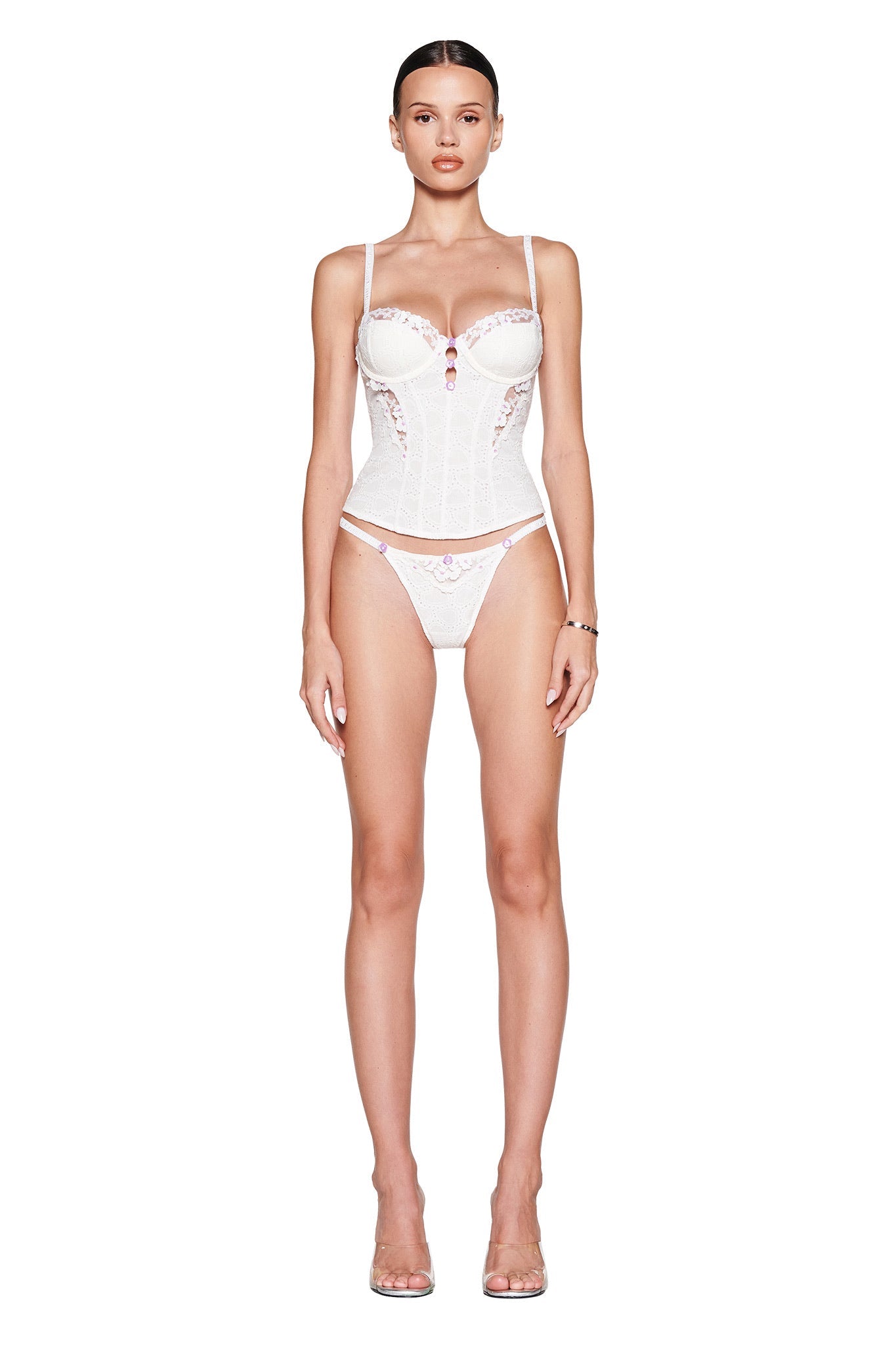A woman faces forward against a white background, wearing the I.AM.GIA ELLA BUSTIER SET in white, featuring a sheer floral tulle bustier with adjustable straps and matching panties. She completes the look with clear heels and a neutral expression.