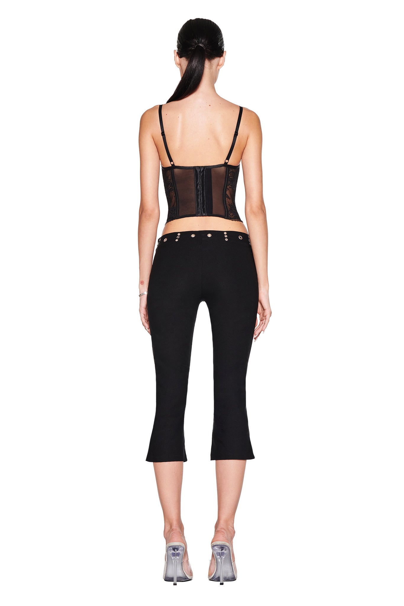 A woman stands with her back to the camera, hair tied back, wearing a black sheer corset-style top, silver heels, and I.AM.GIA SHARAH CAPRI - BLACK: slim black capri pants with a low-rise waistband and grommet details.