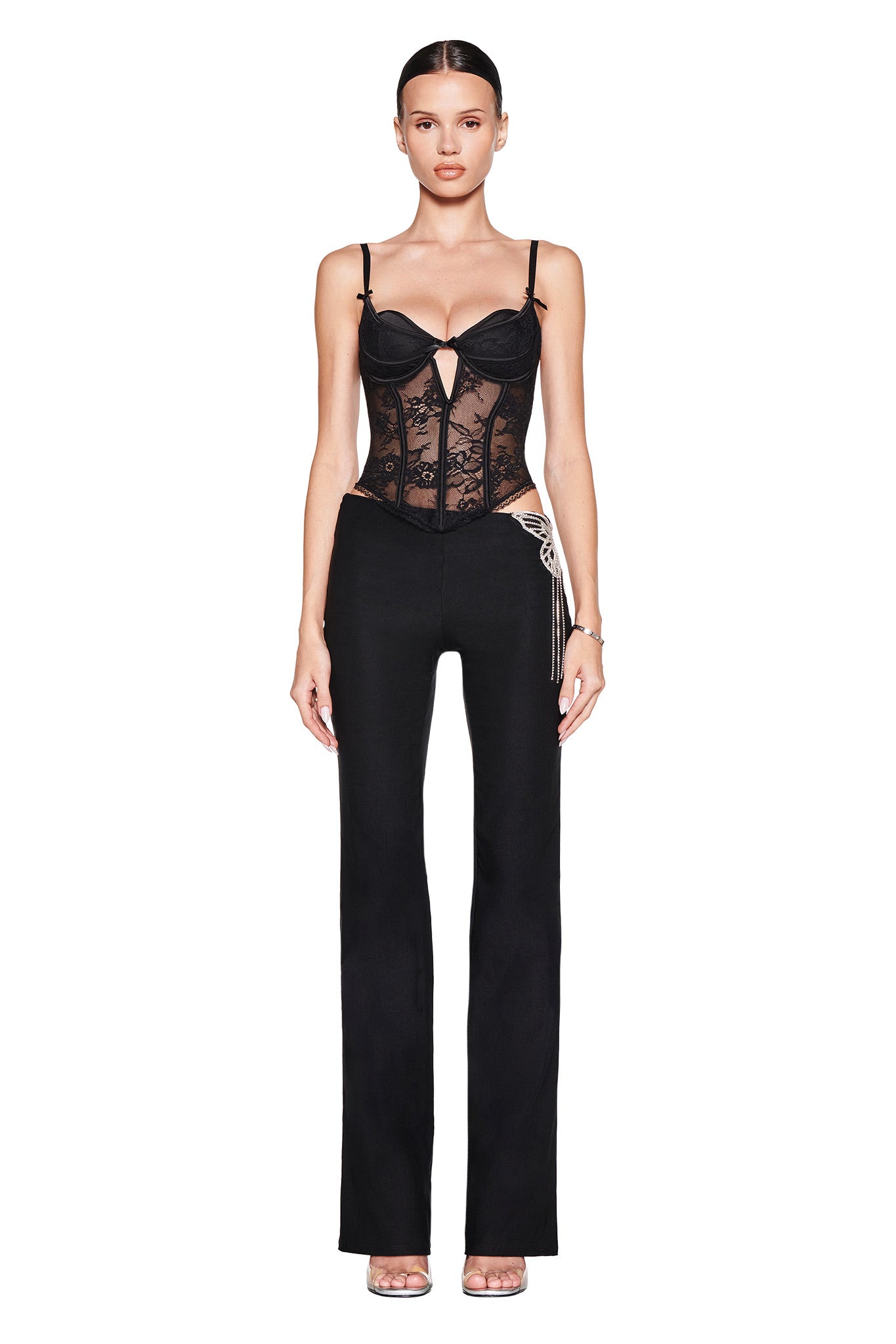 A woman faces forward in I.AM.GIA’s SORREL PANT - BLACK, styled with a black lace corset top featuring an asymmetrical cutout and thin straps. She pairs this look with clear heels, slicked-back hair, and neutral makeup against a white backdrop.